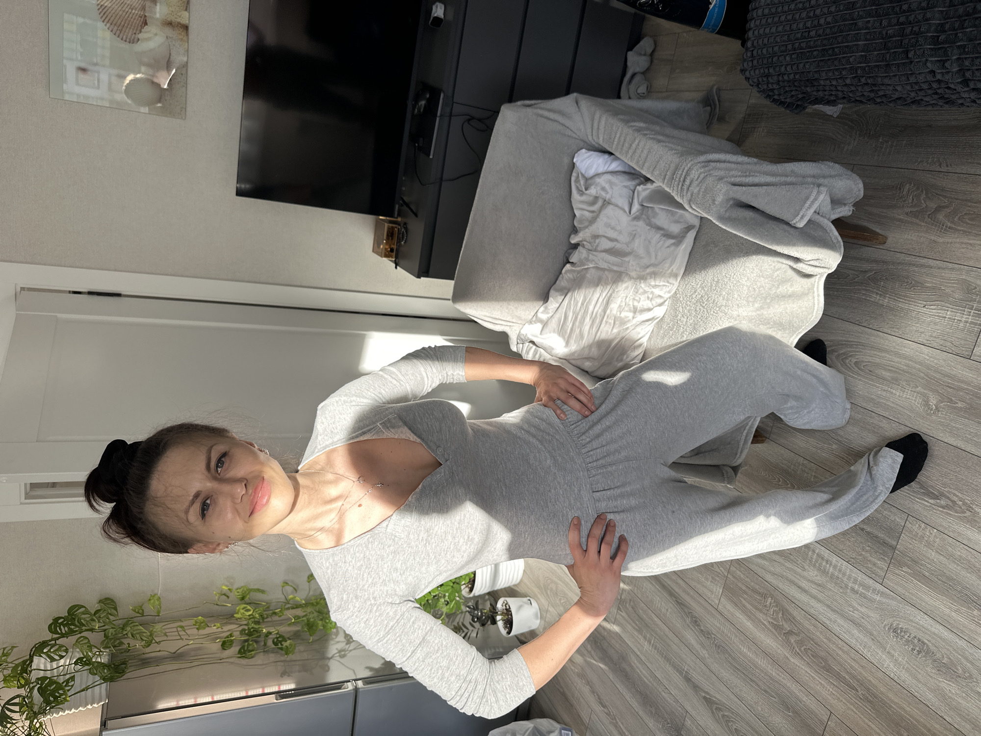 A woman is posing indoors. She is wearing a gray jumpsuit and black socks, with her hair up in a bun. She has her hands on her hips. Behind her is a gray dresser with a TV on top. There is also a gray upholstered chair with a white garment on it. A plant in a white pot is also visible. The background appears to be an interior room with light-colored walls and wood flooring.