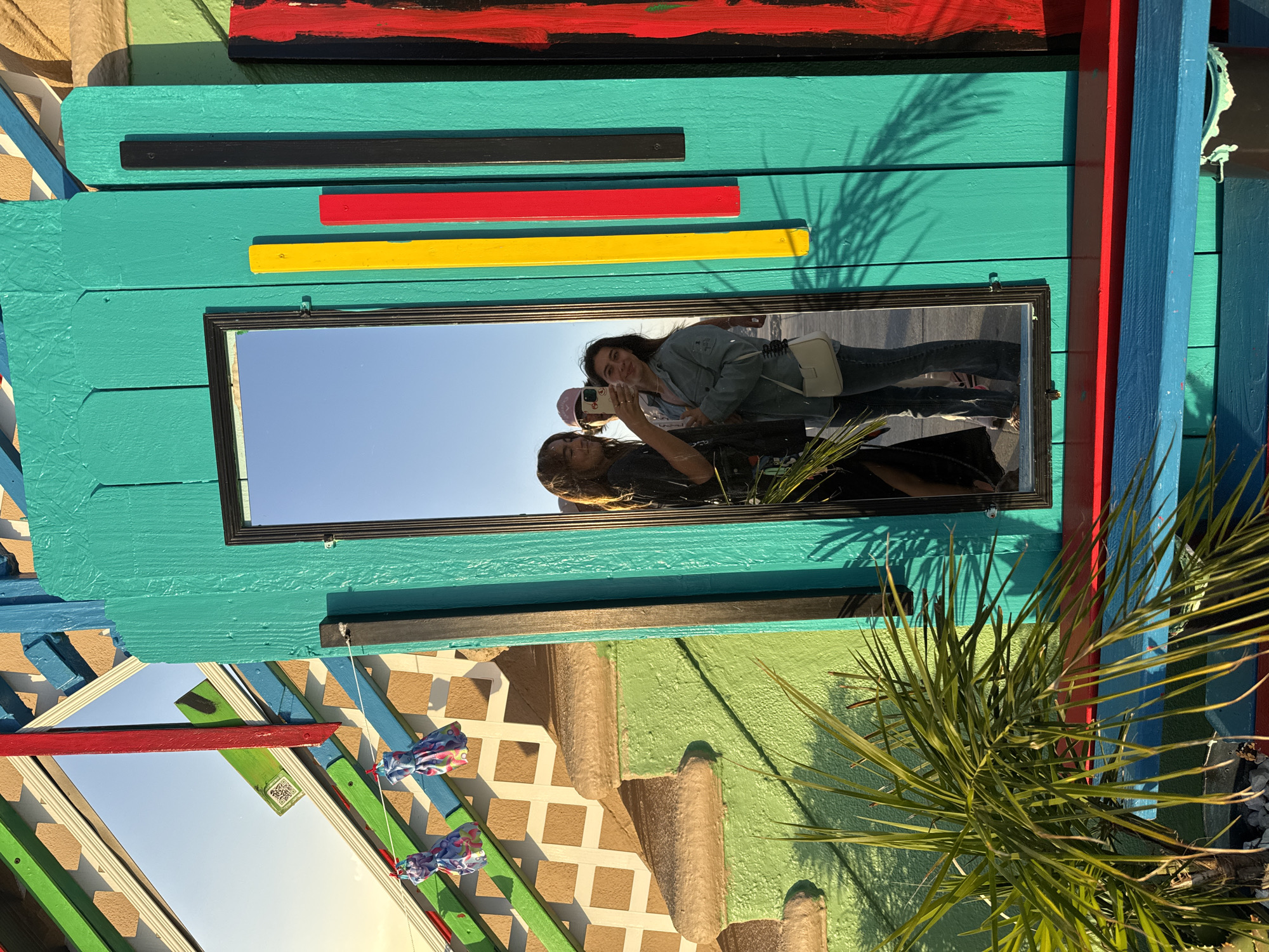 The image shows an outdoor scene in Los Angeles, California, United States. A rectangular mirror, framed in black, is mounted on a teal painted wooden structure with horizontal black, red, and yellow slats. Two people are reflected in the mirror: one is holding a cell phone up in front of their face as if taking a picture, and the other person is standing next to them and looking at the phone. A palm tree is in the foreground, partially obscuring the lower right of the frame. The rest of the wooden structure is painted in bright colors like blue, red, and green, with some elements of tan and white lattice work.