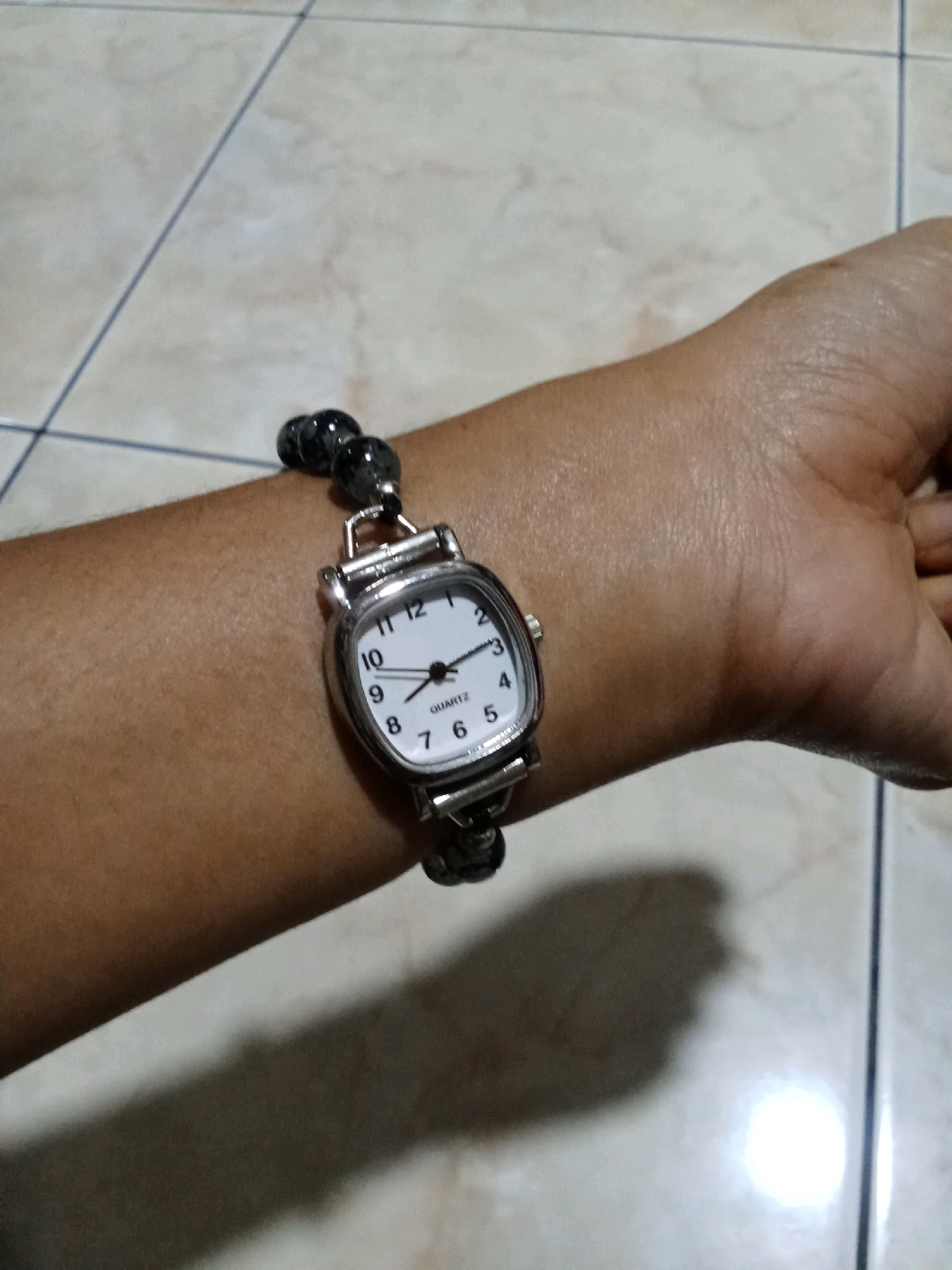 A close-up shot shows a woman's wrist wearing a watch. The watch has a white face with black numerals and hands. It is attached to a beaded bracelet. The background is a tiled floor. The scene is not identifiable as being in West Jakarta, Indonesia.