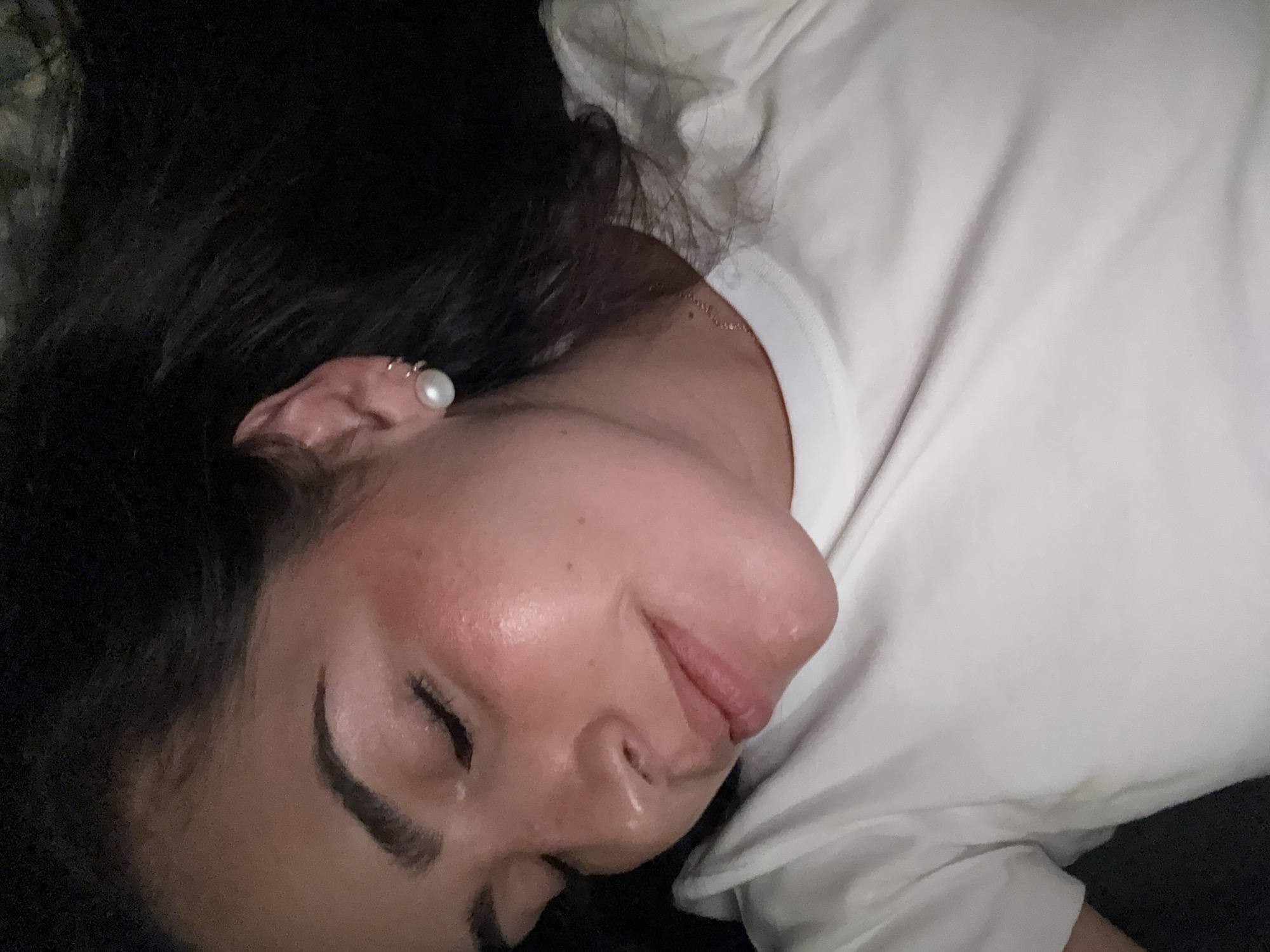 A woman lies on her side, eyes closed, wearing a white t-shirt and a pearl earring in her left ear. Her dark hair surrounds her head. The background is dark and blurry. The location of San Bernardino Tlaxcalancingo, Mexico is not determinable from the image.