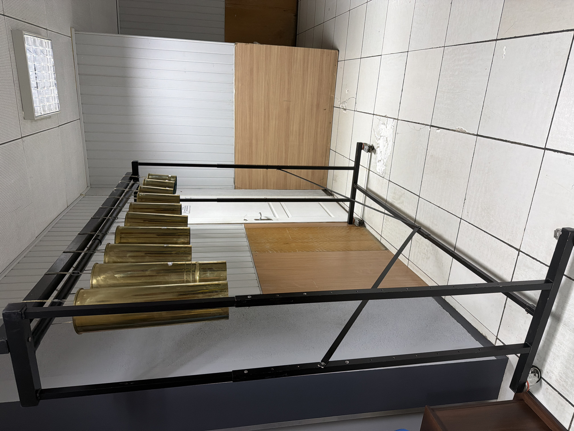 This indoor scene features a black metal structure, resembling a bed frame or display rack, extending from the foreground towards the background. Resting on top of this structure is a row of approximately seven long, golden-colored cylindrical objects, likely made of brass or polished metal. These objects could be chimes, large shell casings, or decorative elements; a small, unreadable white label is visible on one of them.
The floor of the room presents a varied surface. To the right of the metal structure, the floor is covered in light-colored square tiles with dark grout lines, exhibiting noticeable wear, cracks, and patches where the finish is missing. To the left, a section of the floor features light brown wooden panels, adjacent to a light gray textured surface, possibly painted concrete or carpet.
The walls are also composed of different materials. The left wall is covered with light-colored, possibly perforated or grooved panels, featuring a rectangular light fixture with a gridded diffuser. The back wall, situated behind the metal structure, consists of white horizontal paneling. A white door with a simple handle is partially visible on the left side of this back wall. In the far background on the right, a plain wooden board leans against a wall. A small, dark wooden furniture piece, possibly a drawer unit, is partially visible in the bottom right corner.
No people are present, and no specific activity is occurring; the scene depicts a static arrangement. The lighting suggests an indoor environment, likely illuminated artificially. The overall impression is that of a storage, workshop, or display area. No discernible text is clearly visible.