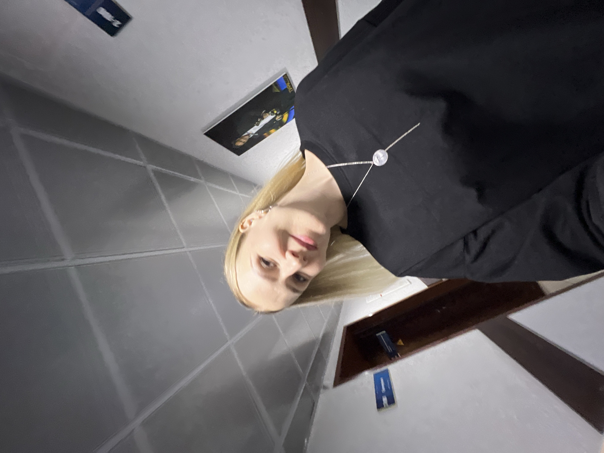 A young woman with blonde hair and fair skin, wearing a black top and a silver necklace, is facing the camera. The background appears to be a room with light grey tiled walls and ceiling. There are framed pictures or signs on the wall. The setting is likely indoors, possibly a bathroom or similar utility space. The location is in Khimki, Russia.