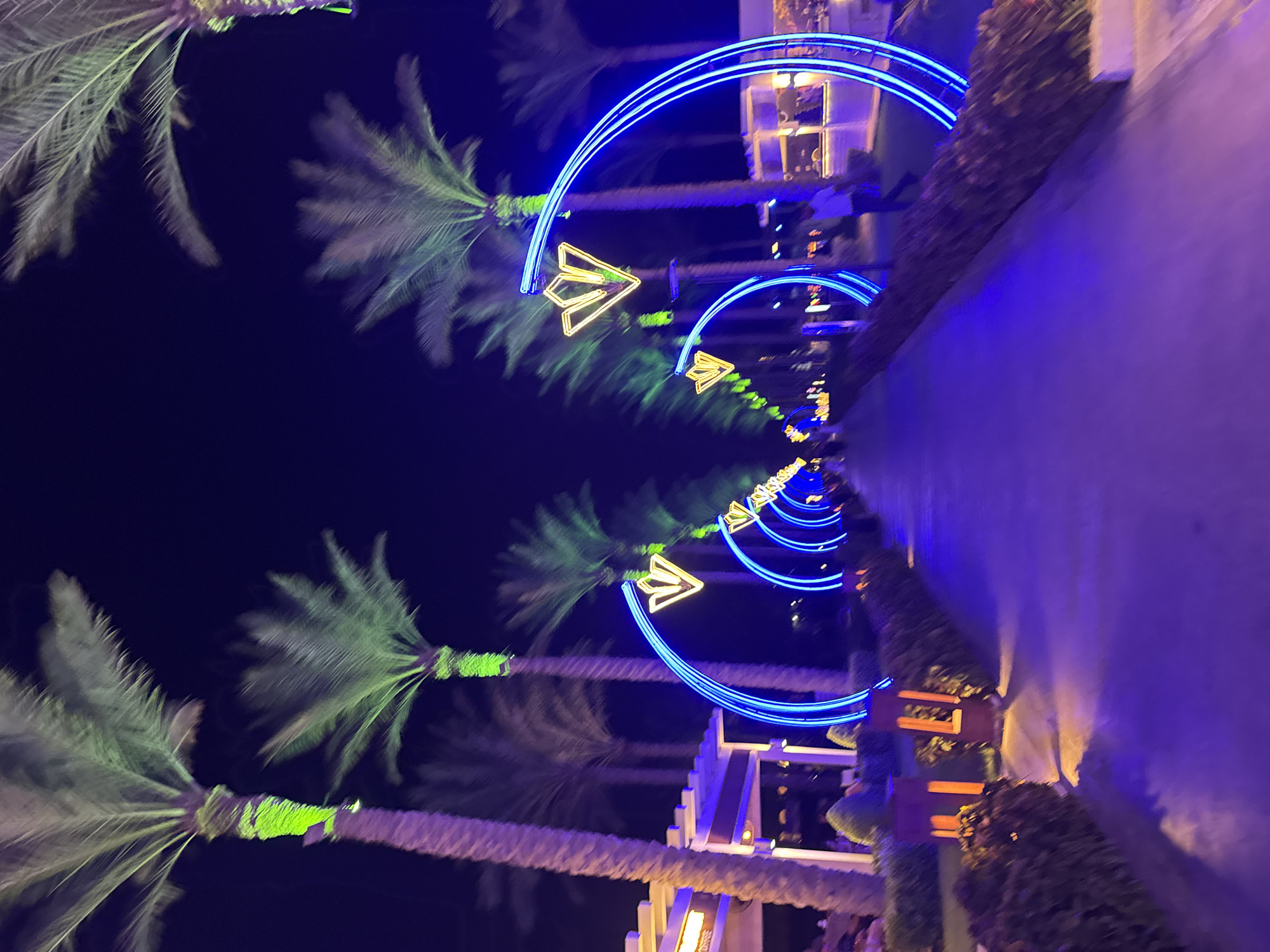 In Unknown, Egypt, this nighttime scene captures a pathway lined with palm trees illuminated by green lights on their trunks. Arched neon blue lights with yellow diamond-shaped accents create a tunnel effect over the walkway. To the left, a building with a white roof and open-air seating is visible. The pathway itself is paved and reflects the blue and purple lights. A few figures are seen walking in the distance. The overall atmosphere is festive and tropical.