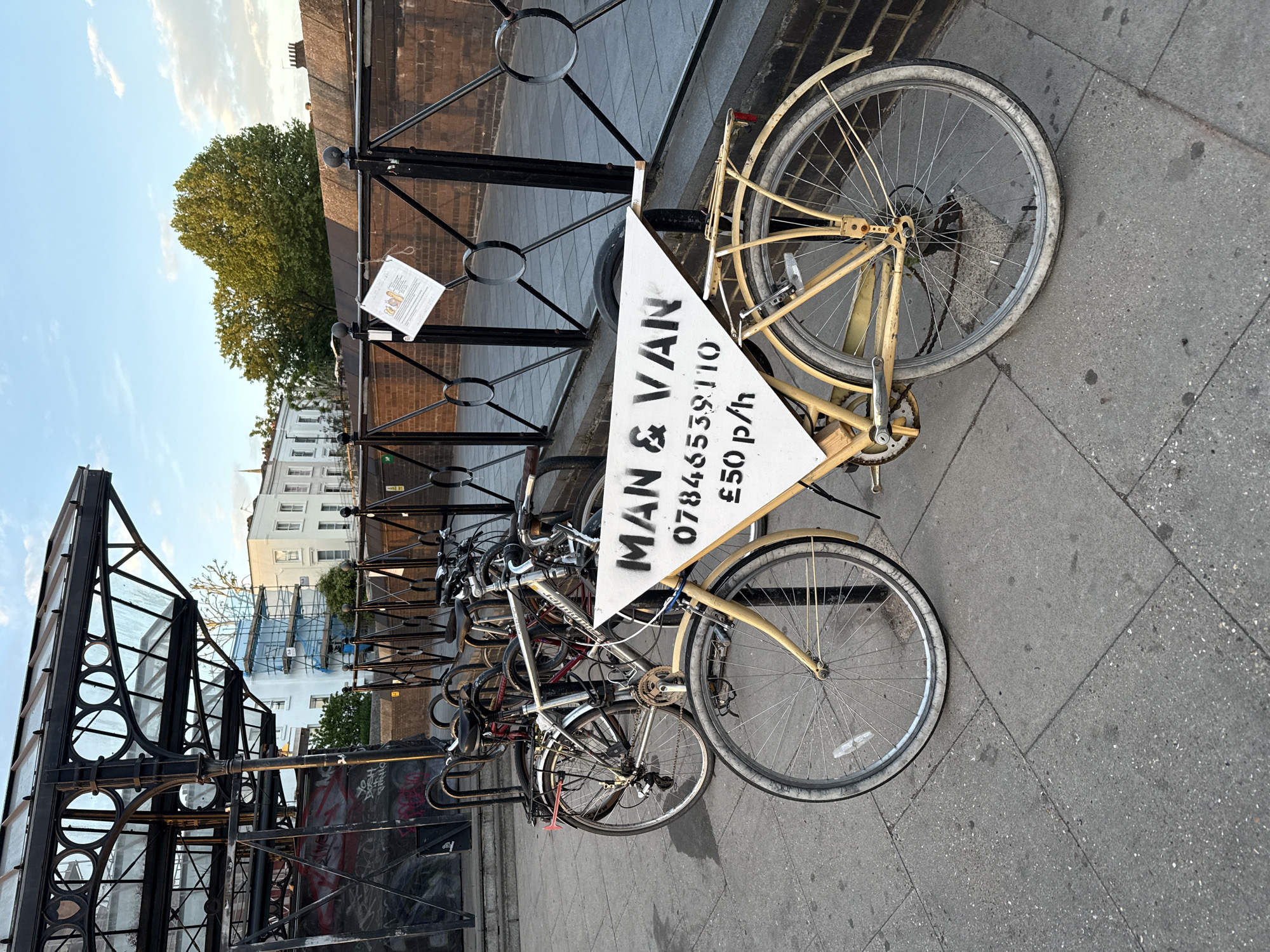 The image shows a row of bicycles parked on a city street, likely in London, United Kingdom. An advertisement for "Man & Van" services is attached to one of the bicycles. The sign displays a phone number (07846539110) and a price (£50 p/h). A structure, possibly the entrance to a subway or other transit station, is visible to the left of the bikes. Buildings can be seen in the background. The street is paved with grey stone. A fence runs along the row of bikes.