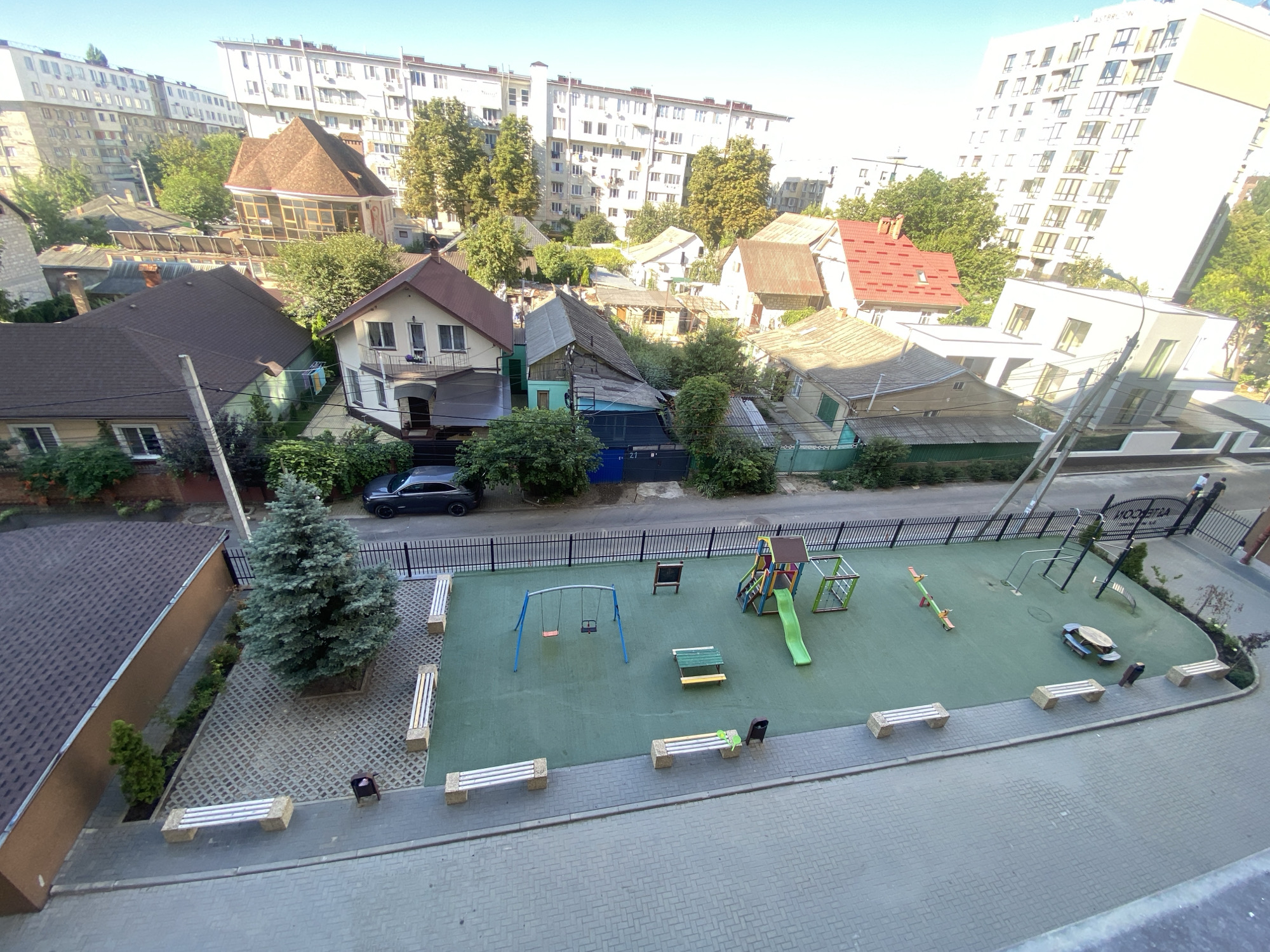 The image shows an aerial view of a residential area in Chișinău, Moldova, on a clear, sunny day. In the foreground, a modern apartment building is partially visible on the left, with its brown tiled roof and tan siding. Adjacent to it is a small, landscaped area with a mature evergreen tree, paving stones, and a few smaller bushes.
The central focus of the image is a children's playground. It features a green rubberized surface with various play structures including a colorful slide and climbing frame, a swing set with two red swings, and a seesaw. Several park benches are placed around the perimeter of the playground, some occupied by a few individuals, suggesting it is a public space. A trash can and a small picnic table are also present within the playground area.
Surrounding the playground are several smaller, older houses with pitched roofs of various materials, including tiles and corrugated metal. The houses exhibit a mix of architectural styles and some show signs of wear. Beyond these houses, larger, more modern apartment buildings dominate the background, with their white facades and numerous windows. Trees are interspersed throughout the scene, adding greenery to the urban landscape. A paved road with a black metal fence runs along the edge of the playground and past some of the houses. A dark-colored car is parked on the side of the road. The sky is a clear, light blue.
