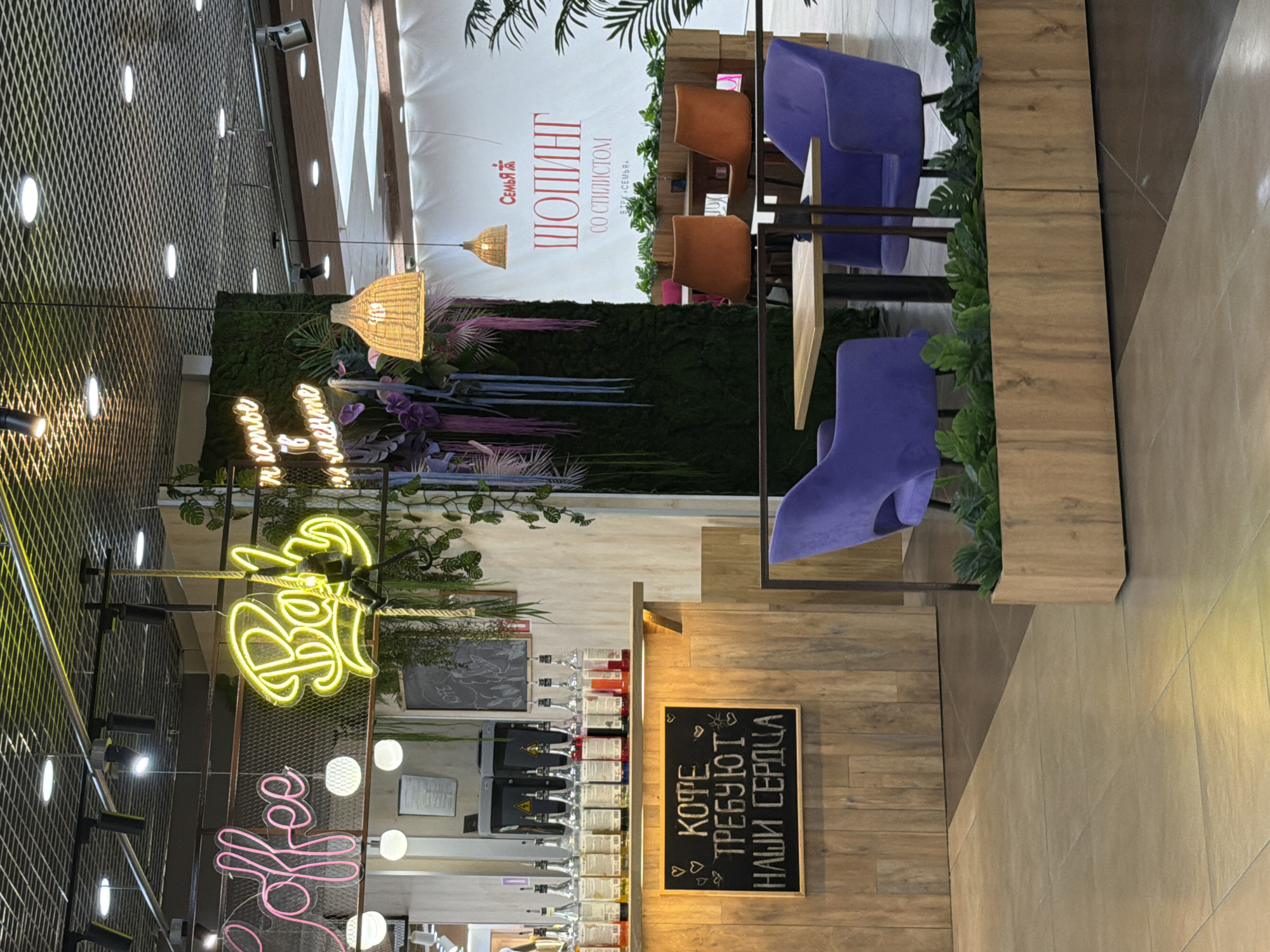 This is a medium shot, taken at a slight upward angle, of an indoor cafe or lounge area, possibly within a shopping mall, given the Russian text that reads "SHOPPING WITH A STYLIST" and "SHOPPING CENTER FAMILY". The overall lighting is warm and inviting, with both ambient and spotlight illumination. The scene features a variety of textures and colors, creating a modern and somewhat eclectic ambiance.
Prominently displayed are two neon signs: one in bright yellow that reads "Coffee" in stylized Russian script, and another in pink, also spelling out "Coffee". These signs are mounted on a metal grid structure that also holds spotlights, suggesting this is likely a cafe or a place serving coffee.
Behind the neon signs, a wall is covered in artificial greenery, creating a striking contrast with the man-made elements. To the right of the greenery wall, there are several modern chairs: two in a vibrant purple and two in a burnt orange color, arranged around small tables. These chairs are set against a backdrop of wooden panels and what appears to be a white banner with Russian text.
Further to the right, a wooden counter or bar area is visible, stocked with various bottles of colorful liquids, possibly syrups or alcoholic beverages. A small chalkboard sign hangs on the wooden wall, displaying handwritten text in white chalk that reads "COFFEE DEMANDS OUR HEARTS". The overall atmosphere suggests a relaxing and stylish spot for patrons to enjoy a beverage and perhaps engage in conversations or shopping activities. The time of day is not discernible, but the indoor setting implies it could be any time.