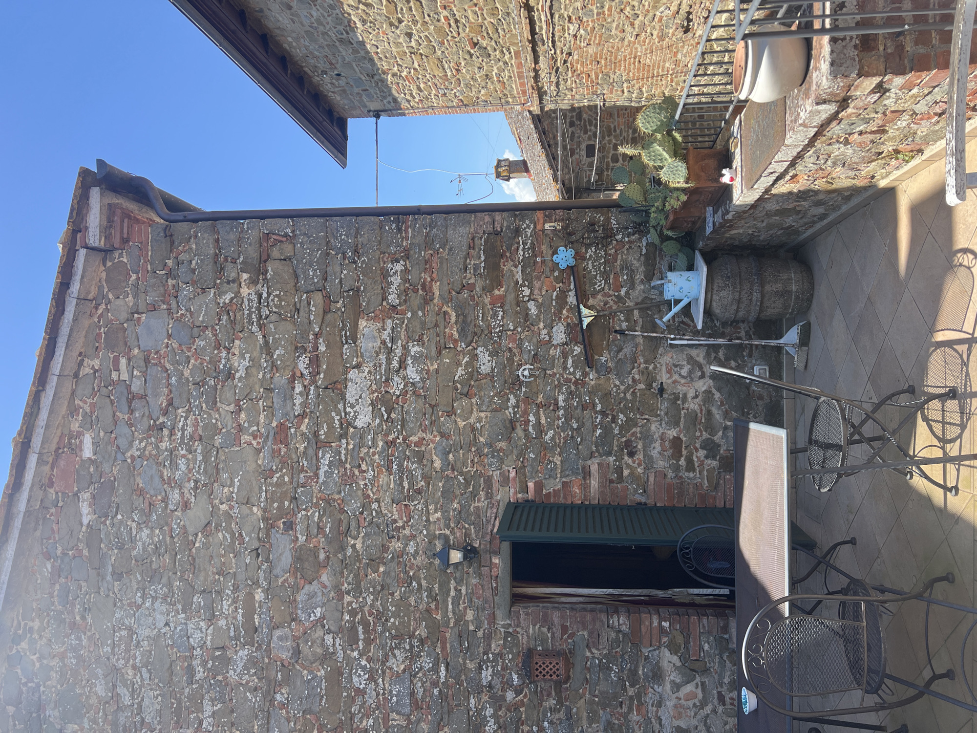 This image captures an outdoor scene in Sinalunga, Italy, centered on the exterior of a stone building under a clear blue sky. The building's facade is constructed with irregularly shaped stones, exhibiting a rough texture and varied coloration of grays and reds. An awning-covered window is visible on the wall, positioned to the side of a light fixture. Various decorative items, including a flower-shaped wall ornament and gardening tools, are affixed to the stone wall. A tiled patio area features a table and metal chairs, bathed in sunlight. Above, a brick structure is partially visible. Adjacent to the patio, a cactus plant is potted near a large barrel.