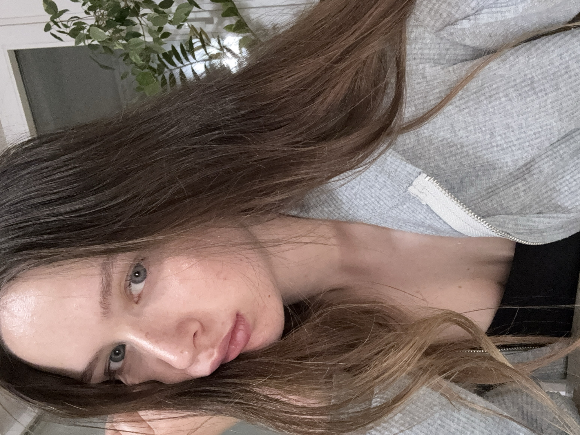 A woman with long brown hair and light blue eyes lies with her head resting on a surface. She is wearing a gray zippered sweatshirt over a black top. The image appears to be taken indoors near a window with foliage visible outside. There is no indication of this being in the city of Chaikovsky, Russia.