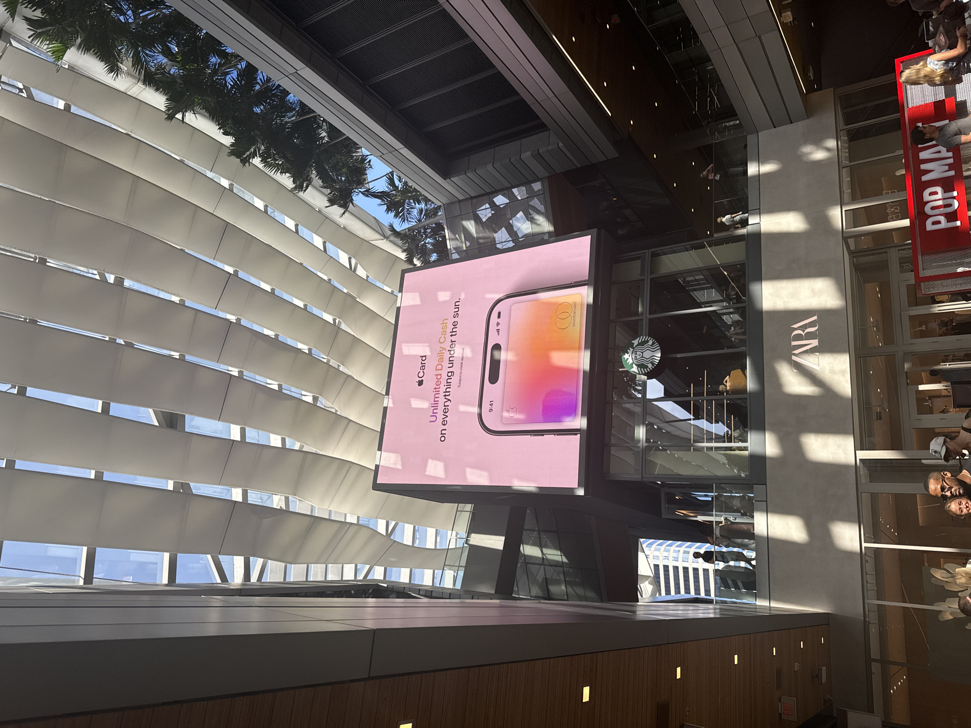 The image captures an architectural scene in the city of Miami, Florida, USA. The camera is oriented upward, revealing a structural combination of exterior and interior elements. A large advertising display featuring an Apple Card promotion is prominently visible, showcasing the card design and slogan "Unlimited Daily Cash on everything under the sun." Adjacent to the advertisement are storefronts for Starbucks, Zara, and Pop Mart. People are seen through the storefront glass, suggesting pedestrian activity. The architecture includes slatted sunshades and glass facades, indicative of modern urban design. Palm trees are visible through the open architectural elements.