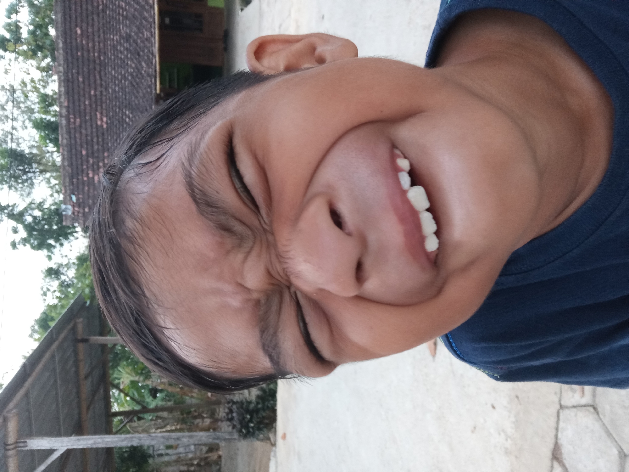 A close-up, low-angle shot captures a young boy making a funny face outdoors. The boy, with dark hair, squints his eyes and bares his teeth in a wide grin, his cheeks puffed out. He is wearing a dark blue t-shirt. In the background, a rustic tiled roof house with wooden window frames is visible. To the left, a simple structure with a corrugated metal roof supported by wooden posts stands under a cloudy sky. The ground appears to be concrete or stone paving. The setting is a rural village, consistent with locations such as Kedunggudel, Indonesia, characterized by its simple architecture and natural surroundings.