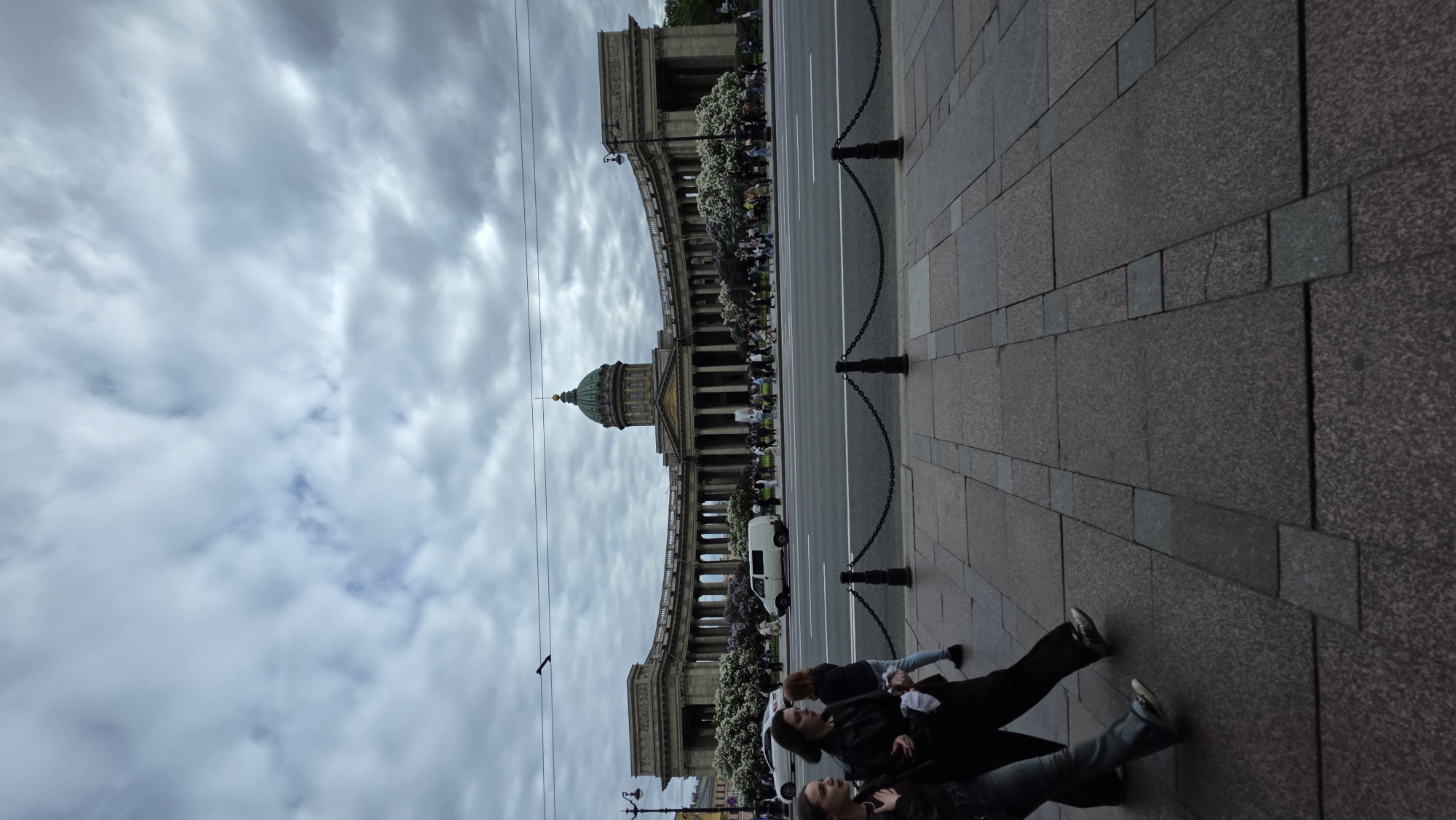 Here is a detailed description of the image:
**Location:**
The image features the Kazan Cathedral in Saint Petersburg, Russia.
**Environment:**
The sky is overcast with a lot of clouds. The sidewalk is paved with granite blocks. There is a street between the cathedral and the sidewalk, and it has a line of black posts connected by chains.
**Subjects:**
There are three people lying on the sidewalk. They are all facing toward the right.
**Objects:**
- Kazan Cathedral
- Granite sidewalk
- Clouds
- Street
- Black posts connected by chains
- Street lamps
- Pedestrians in front of the cathedral
- Van
- Wires