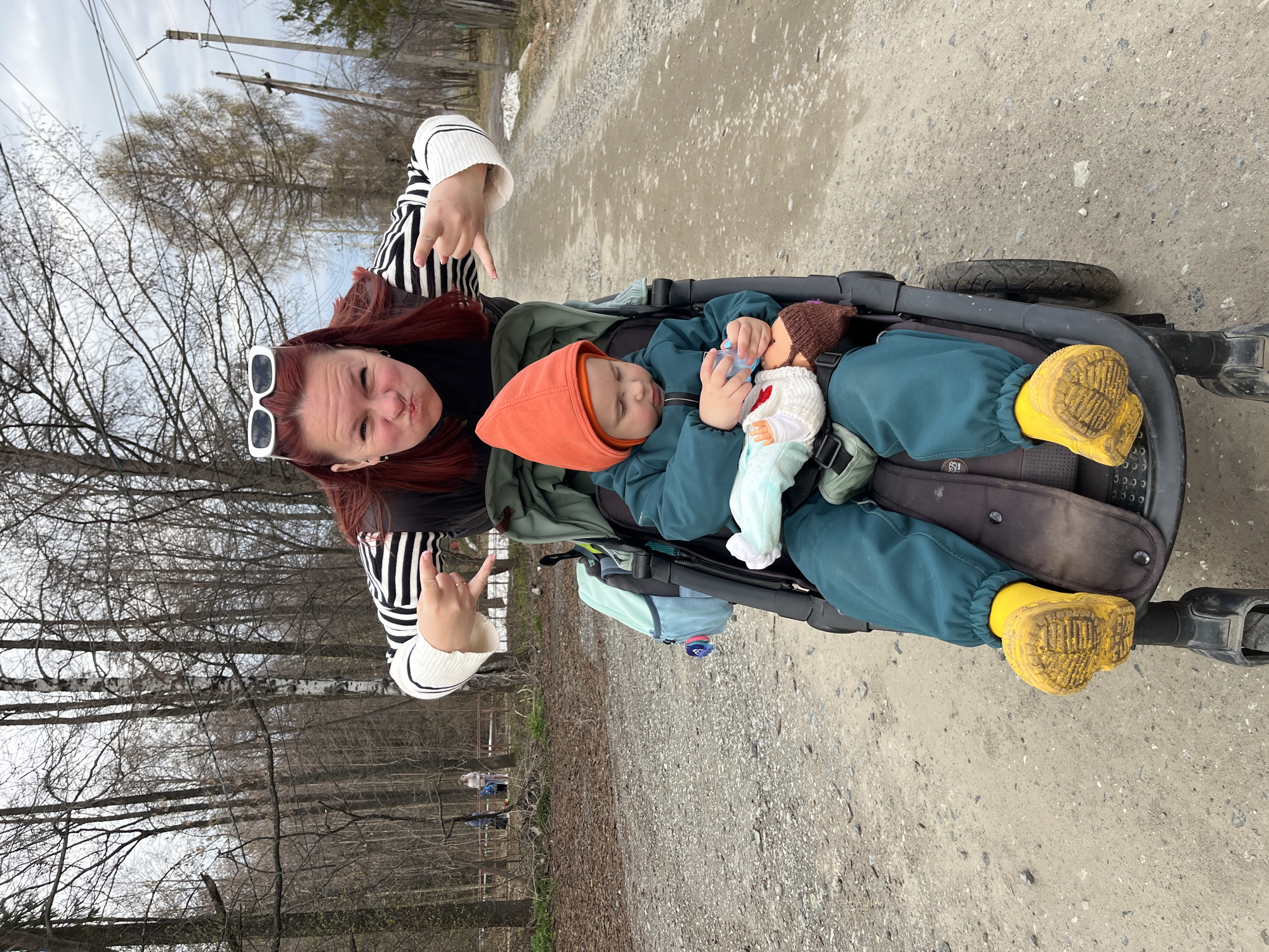 This image, likely captured in Chaikovsky, Russia, features a woman and a child on an outdoor path, surrounded by a sparse natural environment. The scene appears to be set during daytime on an overcast day in late winter or early spring, given the bare trees.
The woman, positioned on the left, has long reddish-brown hair and is wearing white sunglasses pushed up onto her head. She sports a black and white striped long-sleeve shirt under a black top. Her expression is playfully pouting, and she is making "rock on" hand gestures with both hands, conveying a spirited mood.
Beside her, a young child is nestled in a dark-colored stroller. The child is dressed warmly in an orange hooded top, a teal or dark green full-body suit—which has a visible "egeco" tag—and bright yellow boots. The child appears to be resting or asleep, holding a baby bottle up to a doll dressed in white and light blue, which shares the stroller seat. A light blue bag is also attached to the stroller.
The background consists of a gravel or dirt path stretching away, flanked by numerous leafless trees and some distant utility poles with power lines. The overall impression is that of a casual outing or walk in a natural, somewhat rustic setting.