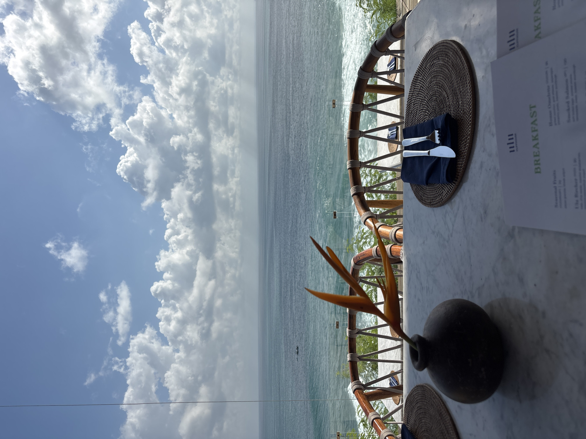 The image shows a table setting on a terrace overlooking the ocean, likely in the Kuta Selatan area of Indonesia, based on the landscape and the "ulu" logo on the breakfast menu which suggests Uluwatu. The sky is predominantly blue with scattered white clouds. The ocean is calm with slight waves. The terrace features a wooden railing with rope accents. On the marble table, there is a place setting with a round woven placemat, a navy blue napkin, and silverware. There is also a dark, round vase with a bird of paradise flower. A menu is partially visible, advertising "Breakfast" and other items. There are no people visible in the image.