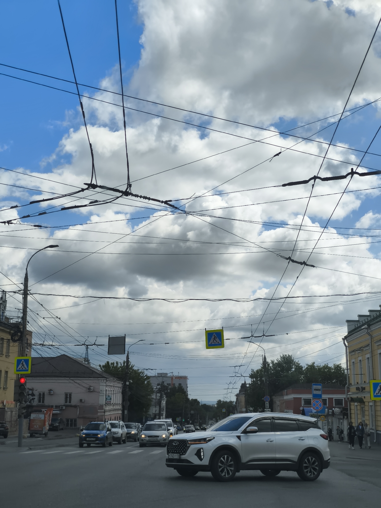 The image captures a vibrant street scene in Izhevsk, Russia, likely at an intersection during the daytime. The sky is partly cloudy with patches of blue, suggesting fair weather. A complex web of overhead power lines, indicative of trolleybus routes common in Russian cities, crisscrosses the upper half of the frame.
The foreground features a white SUV turning or proceeding through the intersection. Several other vehicles, including a blue SUV and white sedans, are visible on the multi-lane road. One traffic light on the left is red. Pedestrian crossing signs are prominent at the intersection, one on the left and another further down the street on the right.
Along the sidewalks, old-style, multi-story buildings with traditional architecture line the street. On the right, a few pedestrians are seen walking. Streetlights and utility poles are interspersed with the buildings and overhead wires. Other road signs include a bus stop sign and a "no stopping/standing" sign.
Visible text includes license plates on the cars: "P742TT 18" on the white SUV, "O204EO 18" on the blue SUV, and "B514BC 18" on another white car, with "18" being the region code for the Udmurt Republic where Izhevsk is located. Some Cyrillic text, possibly "БЕЛОРУС" (BELARUS), is partially visible on a red truck or van on the left side of the street. The scene depicts a typical urban environment with daily traffic and pedestrian activity.