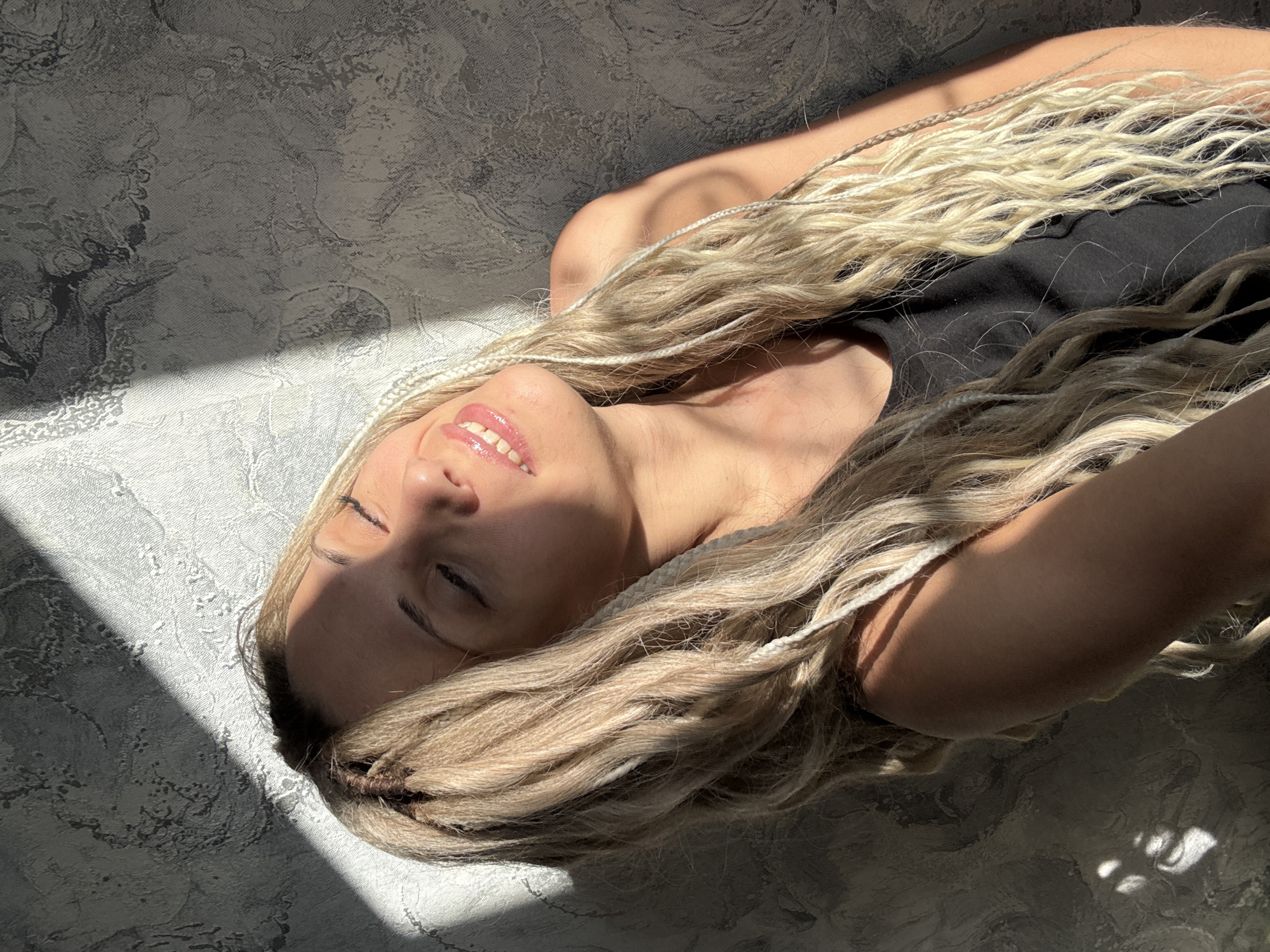 A woman with long, blonde, braided hair is lying on a surface with a grey, textured pattern, possibly a floor or bedding. She is wearing a black top. Sunlight is casting a sharp shadow across her face and the surrounding surface. Her eyes are closed, and she appears relaxed. The setting is unidentified, though the request mentions Rusanovo, Russia.