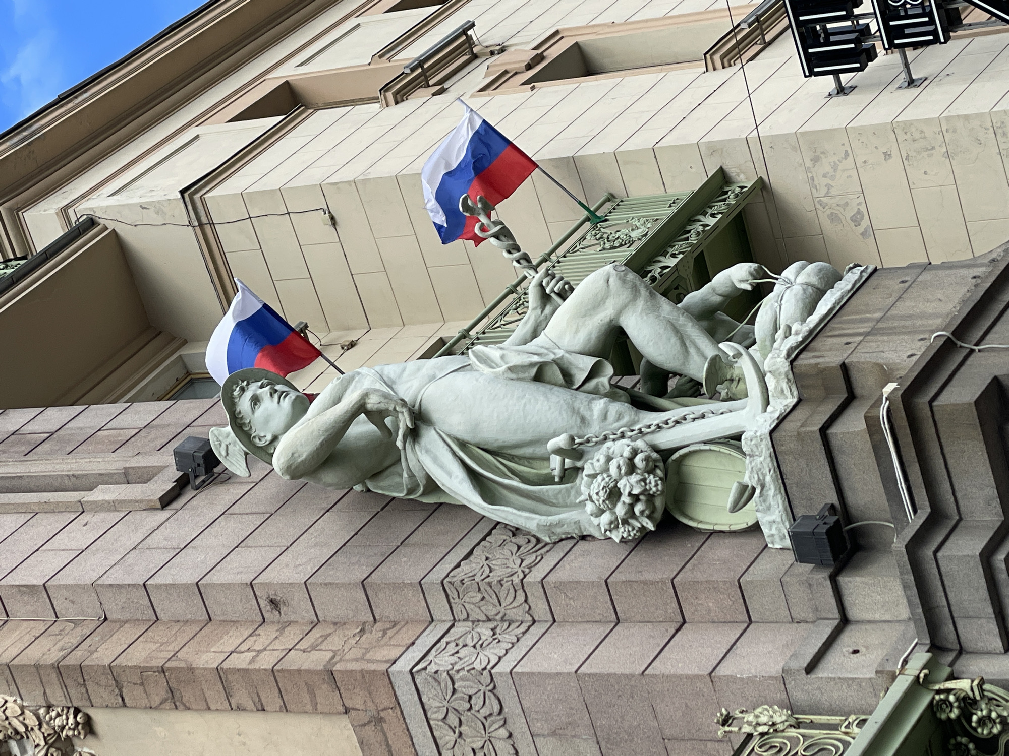 A ground-level view looking up at a building in Saint Petersburg, Russia, features a white statue of a reclining male figure with wings on the side of his head. The statue is holding what appears to be a staff with snakes entwined around it. To the left and behind the statue, two Russian flags are affixed. The statue is positioned on a decorative architectural ledge of the building. The building itself is constructed of light-colored stones or bricks and has several windows and architectural details. A black electrical fixture is visible to the right of the statue. The sky is visible in the upper left corner of the frame.