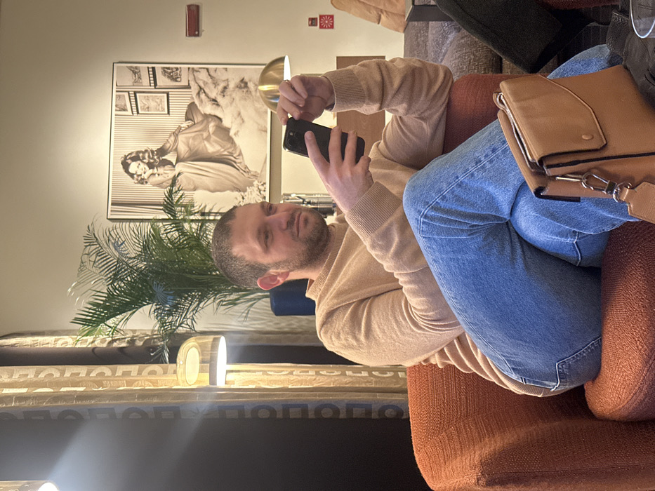A man with short hair, wearing a light brown sweater and blue jeans, sits in an orange chair while looking at his phone. A brown purse is visible next to him. Behind him is a plant and a framed black and white photo. A table with a lamp is in the lower part of the image. The photo is oriented sideways.
