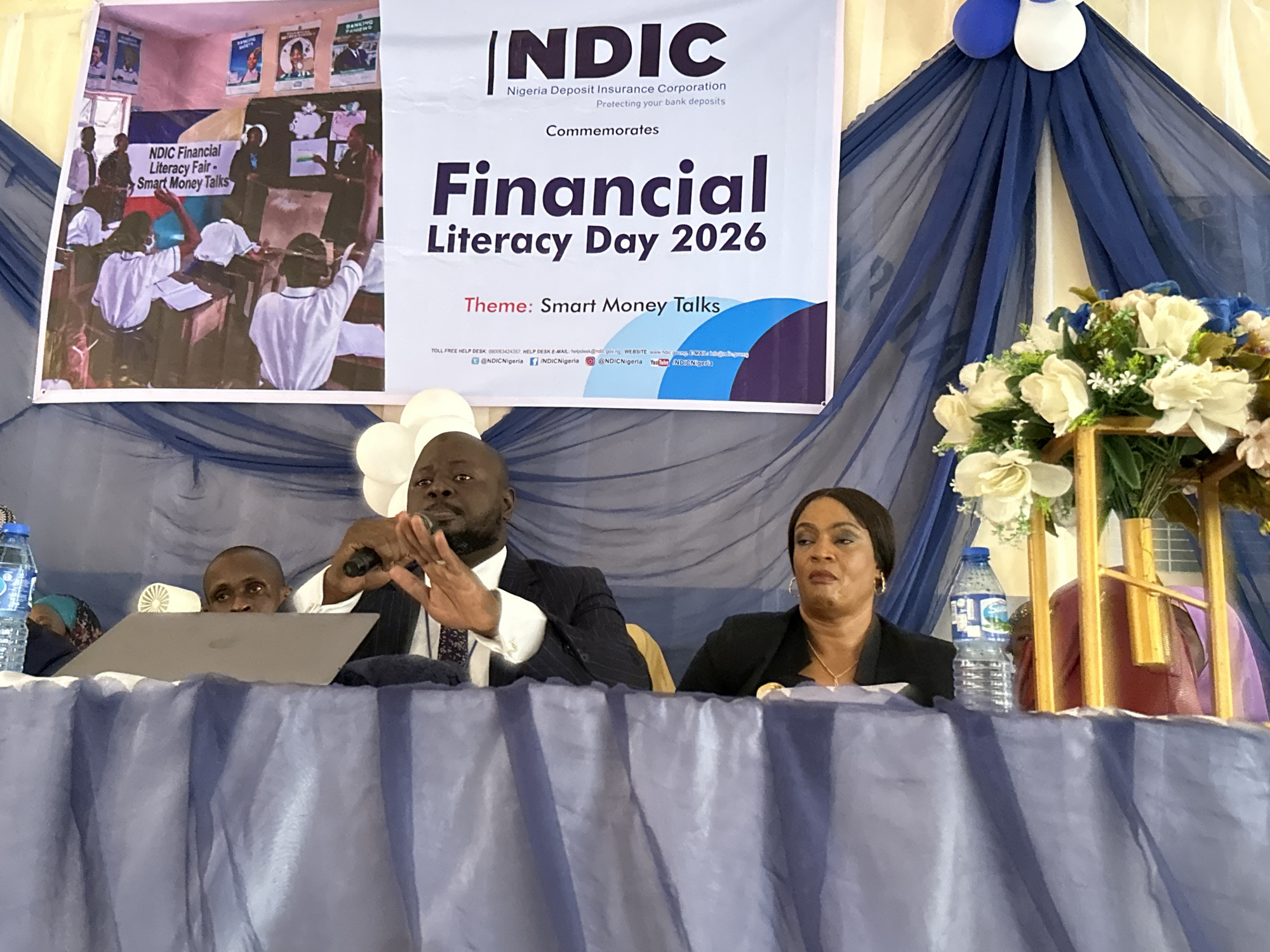 This image captures an indoor event, likely a seminar or conference on financial literacy, held in Unguwan Dosa, Nigeria. The main focus is a large banner in the background that prominently displays "NDIC Nigeria Deposit Insurance Corporation Protecting your bank deposits Commemorates Financial Literacy Day 2026 Theme: Smart Money Talks." An embedded picture on the banner shows a classroom scene, indicating educational activities. Below the event details, contact information including a toll-free help desk number, email, website, and social media handles like "@NDICNigeria" are visible.
In the foreground, two main figures are seated at a blue-draped table. A man, dressed in a suit, is speaking into a microphone, his hands gesturing expressively. To his right, a woman in a dark blazer appears attentive. Another man's head is partially visible behind a laptop to the speaker's left. Water bottles are placed on the table, and a decorative floral arrangement with white and blue flowers sits on the far right. The setting features blue and white drapes and balloons, contributing to a formal atmosphere for the financial education event.
