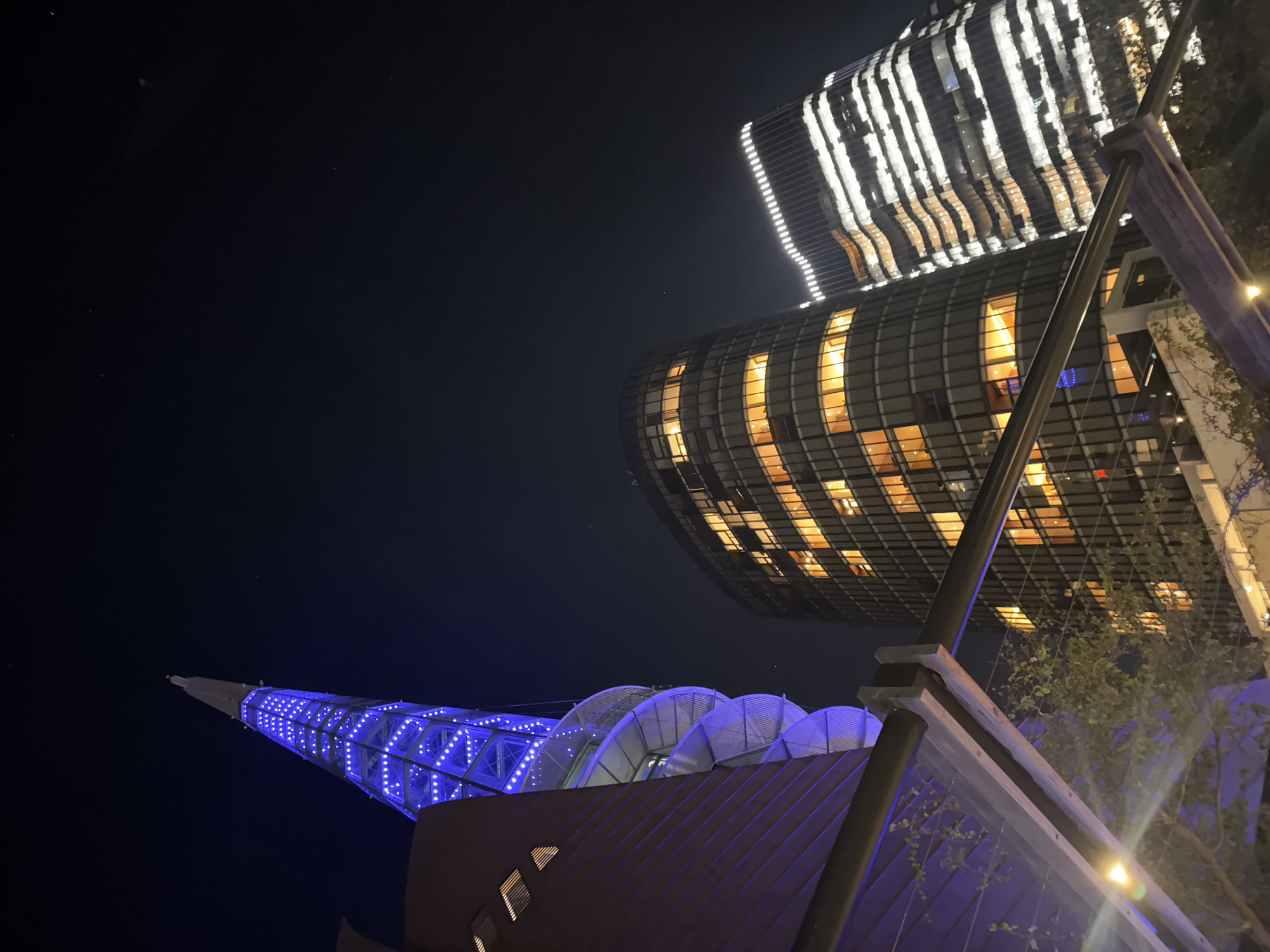 This image captures a striking urban night scene in Perth, Australia, showcasing modern architectural landmarks from a low-angle perspective. The dominant subject on the left is a tall, tapering spire, identifiable as a pylon of the Matagarup Bridge, brightly illuminated with numerous blue LED lights that highlight its lattice-like structure against the dark sky.
Towards the center and right, two contemporary buildings rise into the frame, likely part of the Optus Stadium precinct. The lower and more central building is a large, curved structure with a distinct grid facade, where many windows glow with warm yellow and orange light from within. Above it, partially visible at the top right, is another section of a building characterized by a series of vertical white light strips that create a dynamic, striped pattern.
In the immediate foreground, dark structural poles and some sparse green foliage are visible, framing the illuminated structures. The time of day is clearly night, with a dark, clear sky. The artificial lighting from the buildings and bridge pylon creates a vibrant, modern atmosphere. No people are visible, and there is no discernible text in the image.