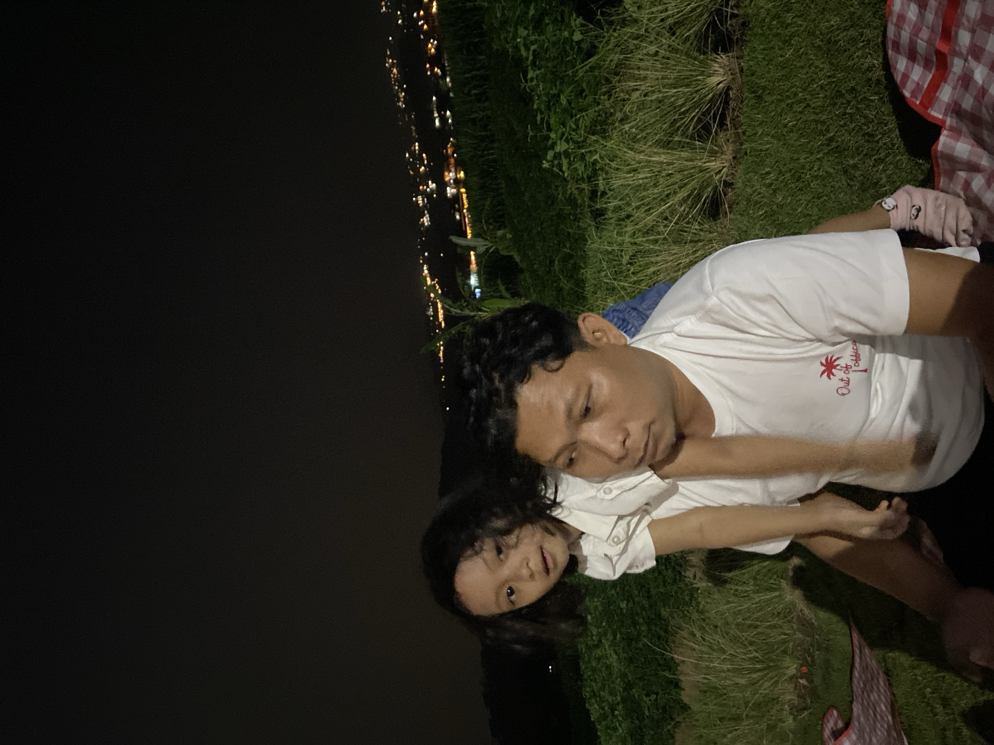 The image shows two people, an adult male and a young child, lying on the grass at night. The adult is wearing a white t-shirt with red text that reads "Out of Office". The child is wearing a white shirt. They appear to be having a moment of closeness. In the background, there are city lights visible in the distance, suggesting an urban or semi-urban setting. The overall scene evokes a sense of peace and quiet, with the dark sky and distant lights creating a serene atmosphere. The presence of a checkered blanket suggests they may be picnicking or relaxing outdoors. The image is taken at night, as indicated by the dark sky and artificial lights. The location is identified as Kesiman, Indonesia.
