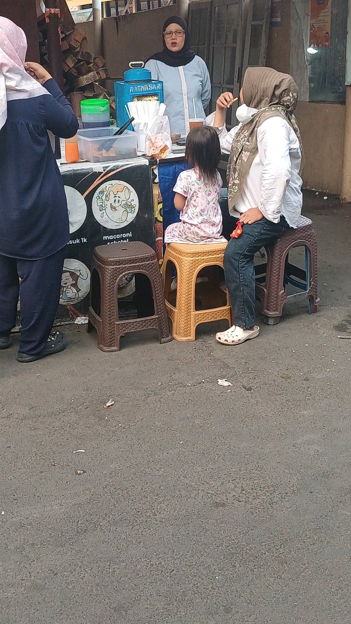 This image captures a street food vendor scene in South Jakarta, Indonesia. A woman wearing a hijab and glasses stands behind a blue food cart labeled "RATNASARI," offering items to customers. To her left, a woman in a dark blue outfit and pink headscarf is partially visible, seemingly interacting with the vendor. In front of the cart, a young girl in a pink and white patterned dress sits on a small yellow plastic stool, facing away from the camera. Another woman, also wearing a hijab and a white top, sits on a brown plastic stool to the right of the girl, eating something. The ground is paved with asphalt, and there are a few discarded wrappers and leaves scattered around. The setting appears to be an outdoor urban environment, likely a sidewalk or street corner.