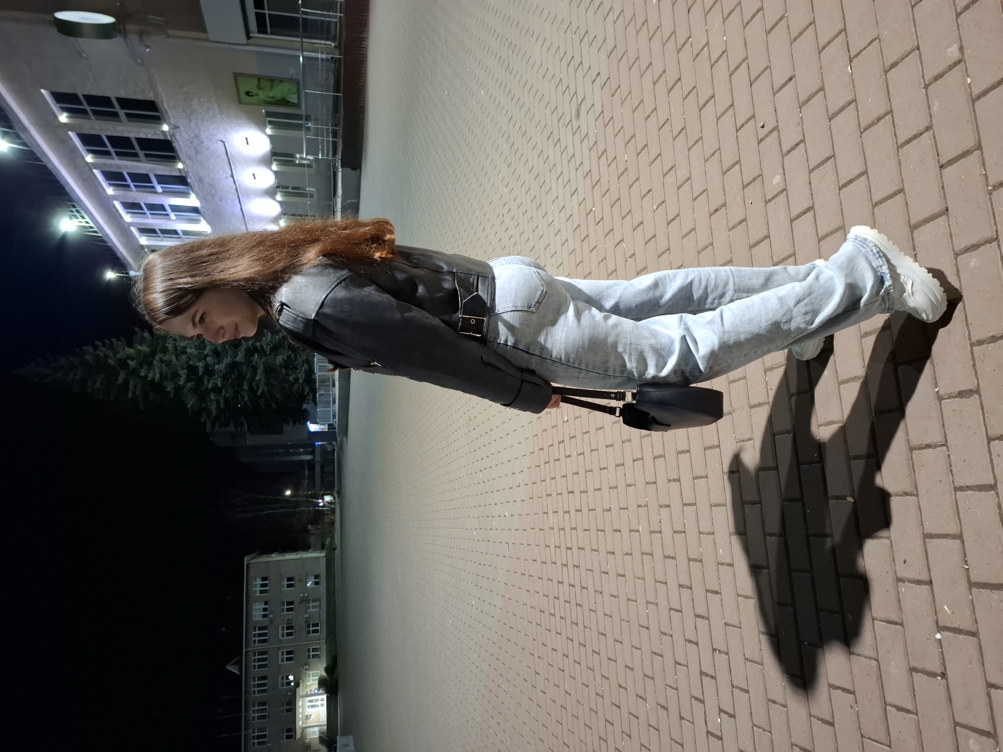 A young woman stands in front of a brick-paved area in the city of Chaikovsky, Russia. She wears a black jacket, light blue jeans, and white sneakers, holding a black bag. The scene appears to be at night, with artificial lighting illuminating the area. Buildings are visible in the background, including one with multiple windows. The woman's shadow stretches across the brick pavement.