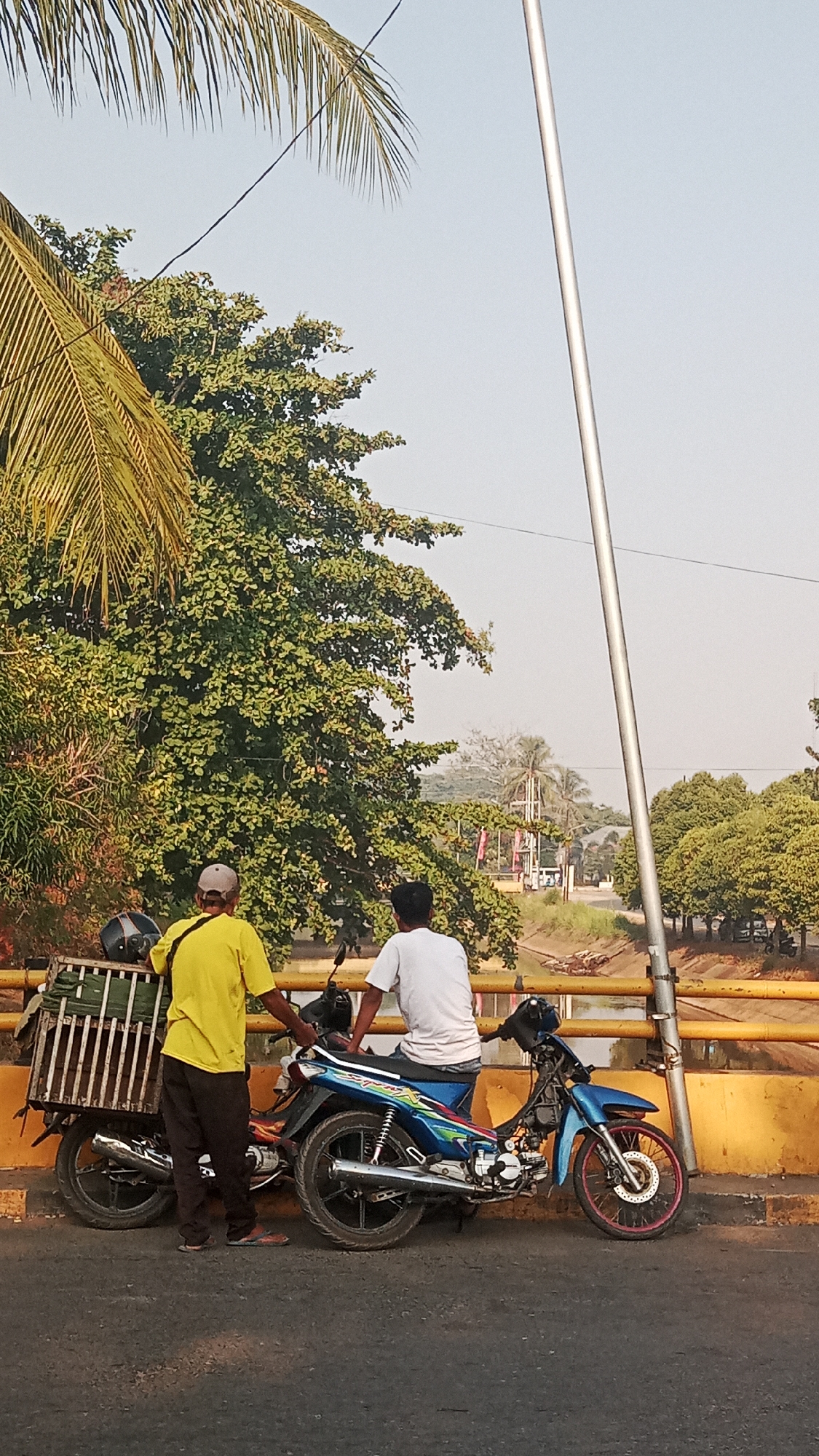 Two men stand on a street next to two motorcycles, one of which is loaded with a cage-like cargo carrier. The man on the left wears a yellow t-shirt and dark pants, and a cap. The man on the right wears a white t-shirt and dark shorts. They appear to be on a bridge with a yellow railing overlooking a body of water. Lush green trees are visible in the background, with a clear, bright sky above. The scene is set in Tanjung Rema, Indonesia.