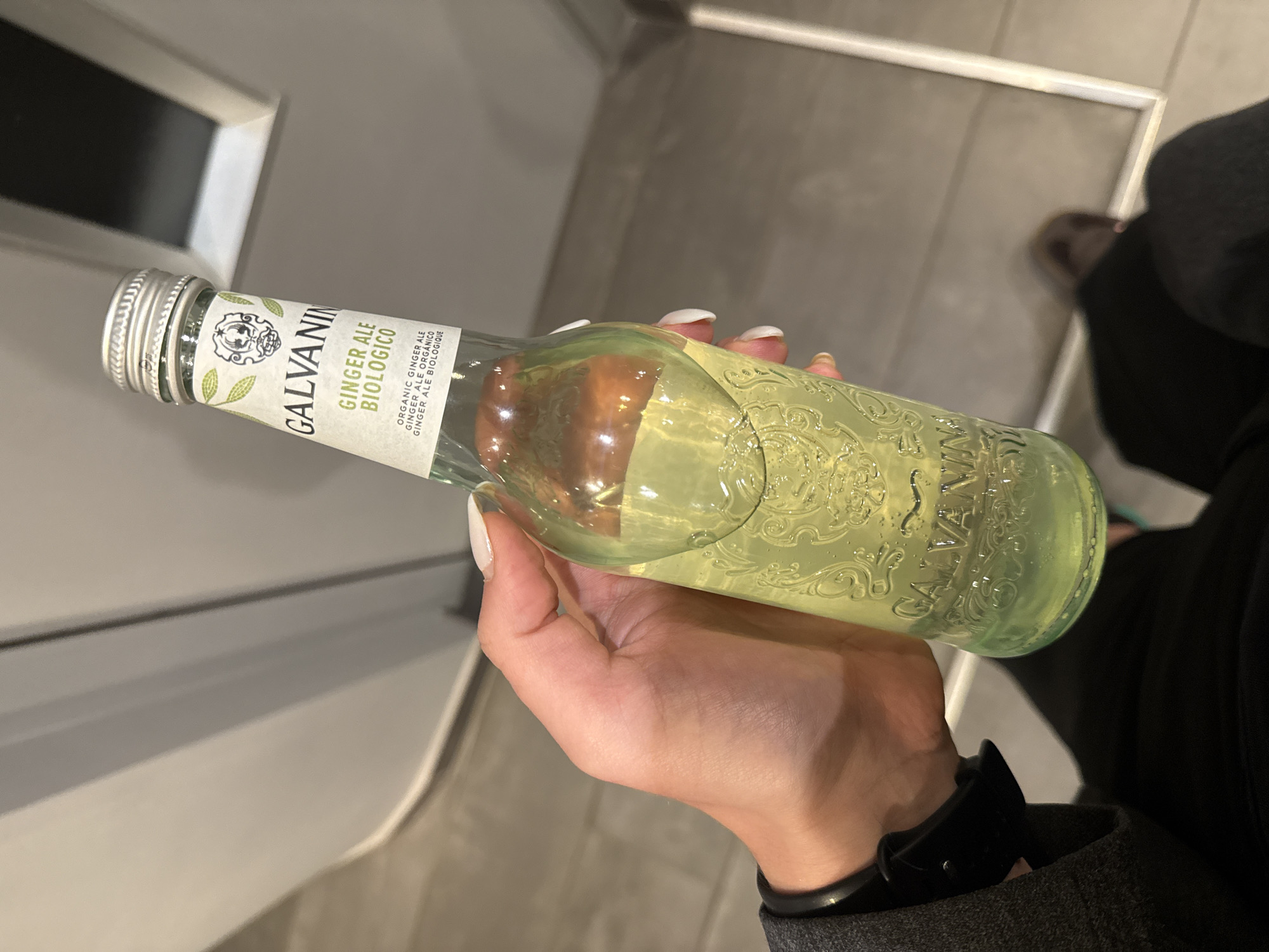 A hand with white manicured nails holds a bottle of Galvanina Ginger Ale Biologico. The bottle is partially filled with a clear, light yellow liquid. The bottle has a silver cap and a white label. The background is an interior setting with a combination of off-white walls and a floor. A portion of a black garment and a wristwatch on the wrist are visible. The location is unknown, and it is impossible to determine if it is in Paris, France.