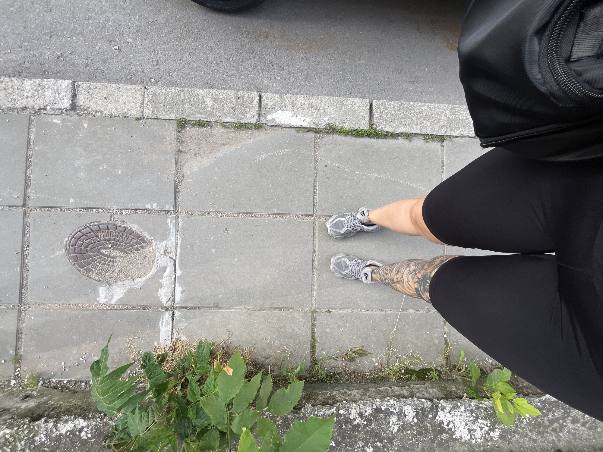 An overhead shot captures a person standing on a concrete sidewalk next to an asphalt road, in what is identified as Veliko Tarnovo, Bulgaria. The person is visible from the upper thighs down, wearing black form-fitting leggings and grey athletic shoes. An intricate dark tattoo covers the lower part of their left leg. A black bag or backpack is partially visible in the upper right corner of the frame.
The sidewalk is constructed from square concrete pavers, with weeds and small patches of grass growing in the cracks and along the concrete curb that separates the sidewalk from the darker asphalt road. To the left, embedded in one of the sidewalk slabs, is a circular metal manhole cover. The cover is partially obscured by dirt and debris but clearly shows the embossed word "HYDRANT" and a radiating pattern. Additional sparse vegetation, including leafy green plants, grows along the edges of the sidewalk and curb in the foreground.
The lighting suggests it is daytime, with no harsh shadows indicating strong direct sunlight, suggesting either overcast conditions or indirect light. The overall scene depicts a typical urban or semi-urban street environment, with common infrastructure elements and some natural growth.