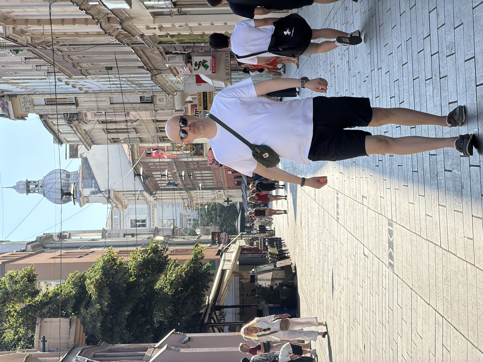 The image depicts a street scene in Sarajevo, Bosnia and Herzegovina, characterized by narrow, pedestrianized cobblestone pathways lined with historic buildings. In the foreground, a bald man wearing sunglasses, a white t-shirt, and black shorts walks towards the camera, carrying a green crossbody bag. Behind him, another man in a white t-shirt and grey shorts walks away, with a black messenger bag. Several other pedestrians are visible in the background, some walking, some standing near storefronts. The architecture features a mix of classical and more modern styles, with balconies, awnings, and signage indicating commercial activity. A prominent dome structure with a spire rises in the distance, a characteristic element of Sarajevo's skyline. Sunlight casts long shadows across the street, suggesting it is either early morning or late afternoon. The atmosphere appears to be one of a bustling urban environment on a clear day.