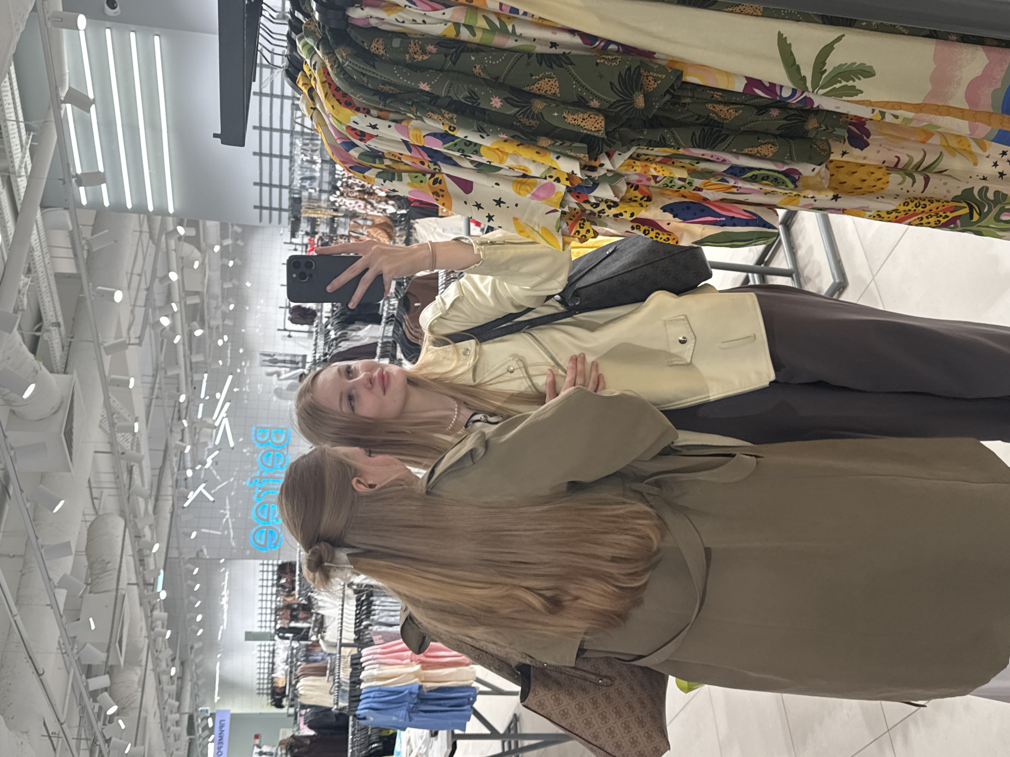 Two young women are taking a mirror selfie in a clothing store. One woman, on the right, is holding up a smartphone and smiling. She is wearing a light-colored jacket and dark pants. The other woman, on the left, is wearing a tan trench coat and has her back to the camera. She is carrying a large tote bag. They are standing in front of racks of colorful clothing. The store appears to be well-lit with fluorescent lighting and has a modern design. There are shelves and displays in the background, indicating it is a retail environment. The reflection in the mirror shows more of the store's interior, including clothing racks and signage that appears to say "Sisters" in blue neon letters. This store is located in the city of Perm, Russia, as indicated by the "Perm" signage visible in the background.