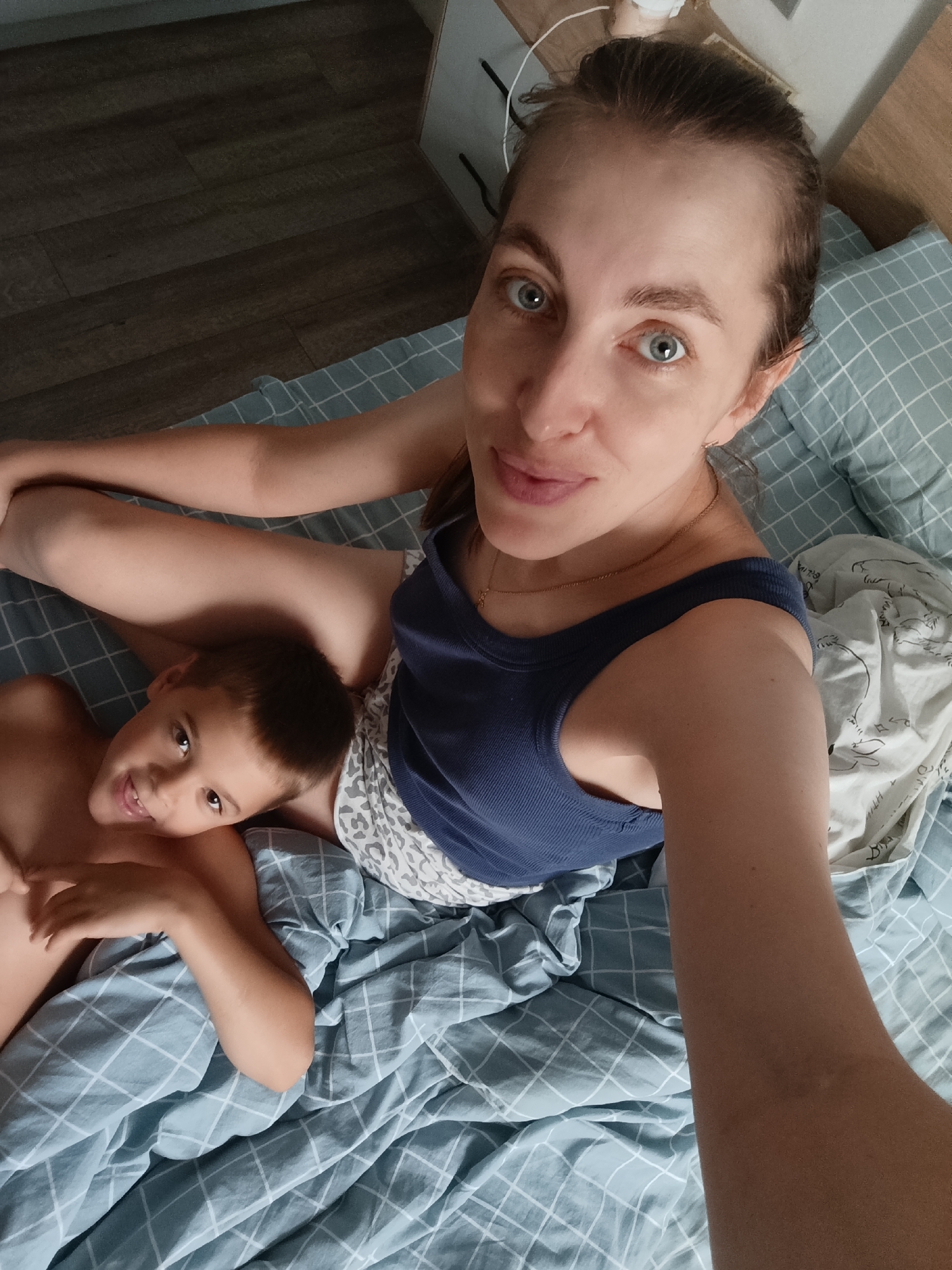 In this selfie, a woman and a young boy are lying on a bed in what appears to be a bedroom. The woman is in the foreground, looking directly at the camera with a slight smile and her eyes wide open. She has her hair pulled back in a bun and is wearing a blue tank top and patterned shorts. Her left arm is bent, resting on her leg, and her right arm is extended as if taking the picture. The boy is positioned to her left, with his head resting on her leg. He is looking towards the camera with a wide smile. He is shirtless. The bed is covered with a blue and white checkered duvet. In the background, a wooden floor is visible, suggesting a domestic setting. The lighting suggests it might be daytime. The overall mood is relaxed and affectionate, indicating a close bond between the mother and son. The location is Chaikovsky, Russia. There is no visible text in the image.