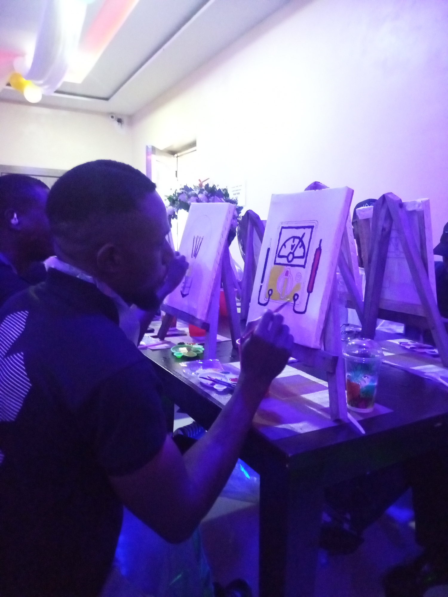 In a brightly lit room with purple and white ambient lighting, two individuals are engaged in a painting activity. The primary subject is a man in the foreground, seen from the side, wearing a dark t-shirt with a white geometric pattern on the left sleeve. He is focused on a canvas placed on a wooden easel, actively painting with a brush. The canvas displays a drawing of a meter with two red and black probes connected. To his left, another person, partially visible, is also attending to their canvas. Several other easels with blank canvases or partially completed paintings are visible in the background. A table in front of the man holds art supplies, including a palette with colorful paints and a clear plastic cup containing a colorful liquid. The overall scene suggests a group painting event or class. The event is taking place at the Fountain Hotel, Ado Ekiti, Nigeria.