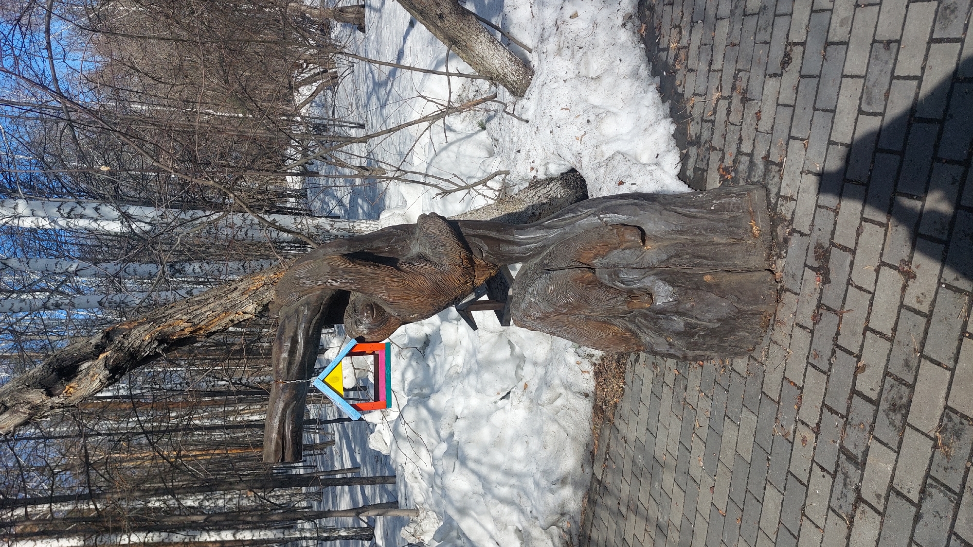 The image shows a wooden sculpture of two bears in a park setting in Novosibirsk, Russia. The sculpture is located beside a paved walkway, with snow present in the background, along with bare trees. A colorful birdhouse is affixed to the sculpture.
