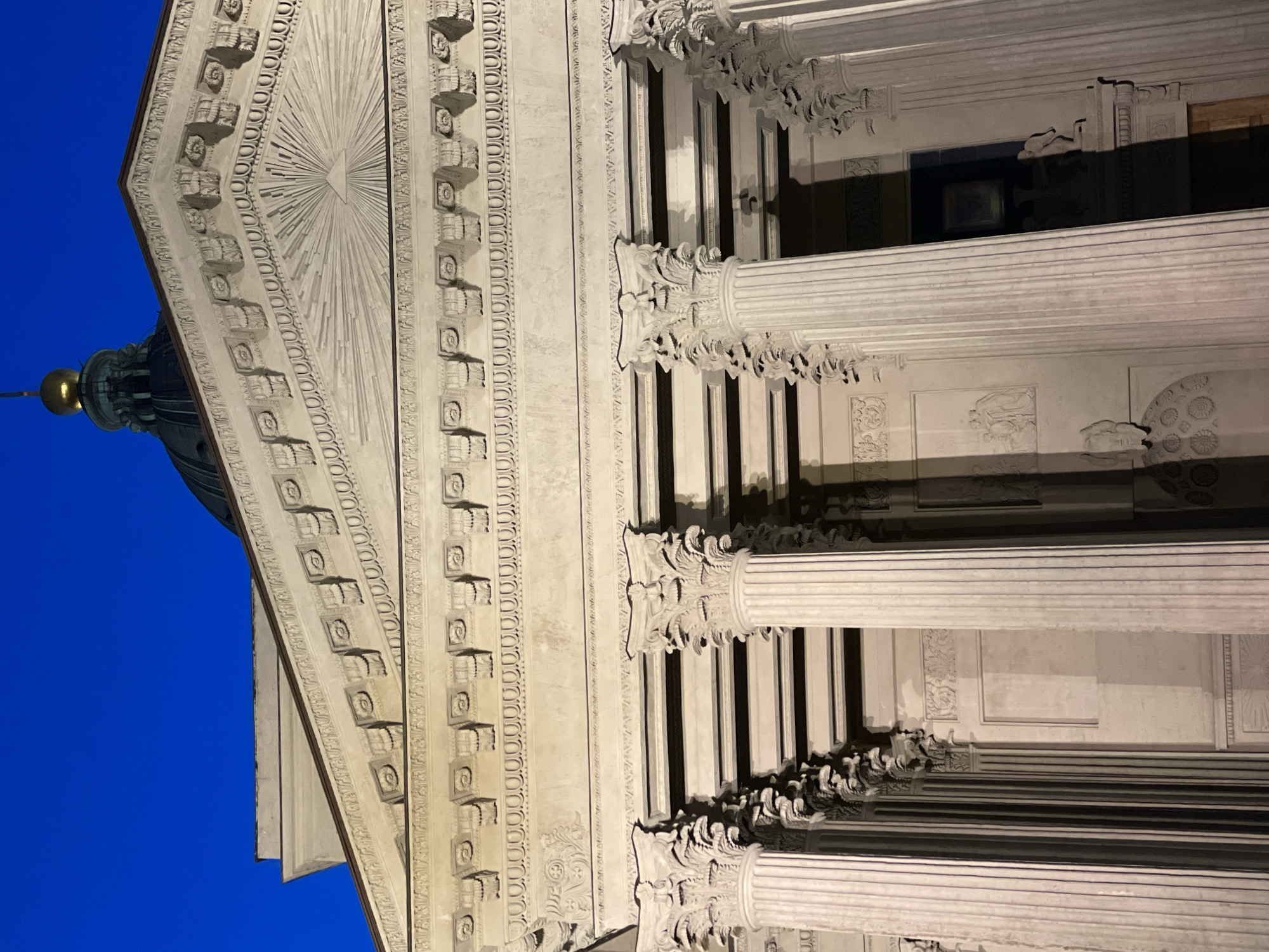 This detailed image captures a close-up, upward-angled view of the Neoclassical facade of the Kazan Cathedral (Kazansky Cathedral) in Saint Petersburg, Russia, observed during twilight or nighttime conditions. The deep blue sky occupies the upper portion of the frame.
The central and dominant feature is the building's entablature and pediment. The triangular pediment, constructed of light-colored stone, features a prominent bas-relief in its tympanum: the "Eye of Providence" (also known as the All-Seeing Eye), represented as an eye within a triangle, from which numerous straight rays emanate outwards.
Below the pediment, the entablature is elaborately decorated with multiple horizontal friezes. From top to bottom, these include a series of small, square-shaped blocks (dentils) directly beneath the pediment's raking cornice, followed by a band featuring regularly spaced, stylized rosettes or circular motifs within recessed squares. Below this, another band displays a continuous pattern of interconnected scrolls and foliage. Further down, directly above the columns, is a frieze with what appears to be an egg-and-dart or bead-and-reel molding.
Several tall, fluted columns with ornate Corinthian capitals are visible, extending vertically from the lower part of the frame. The capitals are richly detailed with sculpted acanthus leaves. The columns cast pronounced vertical shadows, suggesting artificial illumination from the front. Behind these columns, the building's wall features decorative elements, including rectangular panels and bas-reliefs depicting figures or scenes, such as a kneeling figure visible to the far right.
In the upper right corner, against the dark blue sky, the lower portion of the cathedral's large, dark-hued dome is visible, topped by a golden finial. The overall scene conveys the grandeur and intricate architectural detail of this significant Saint Petersburg landmark.