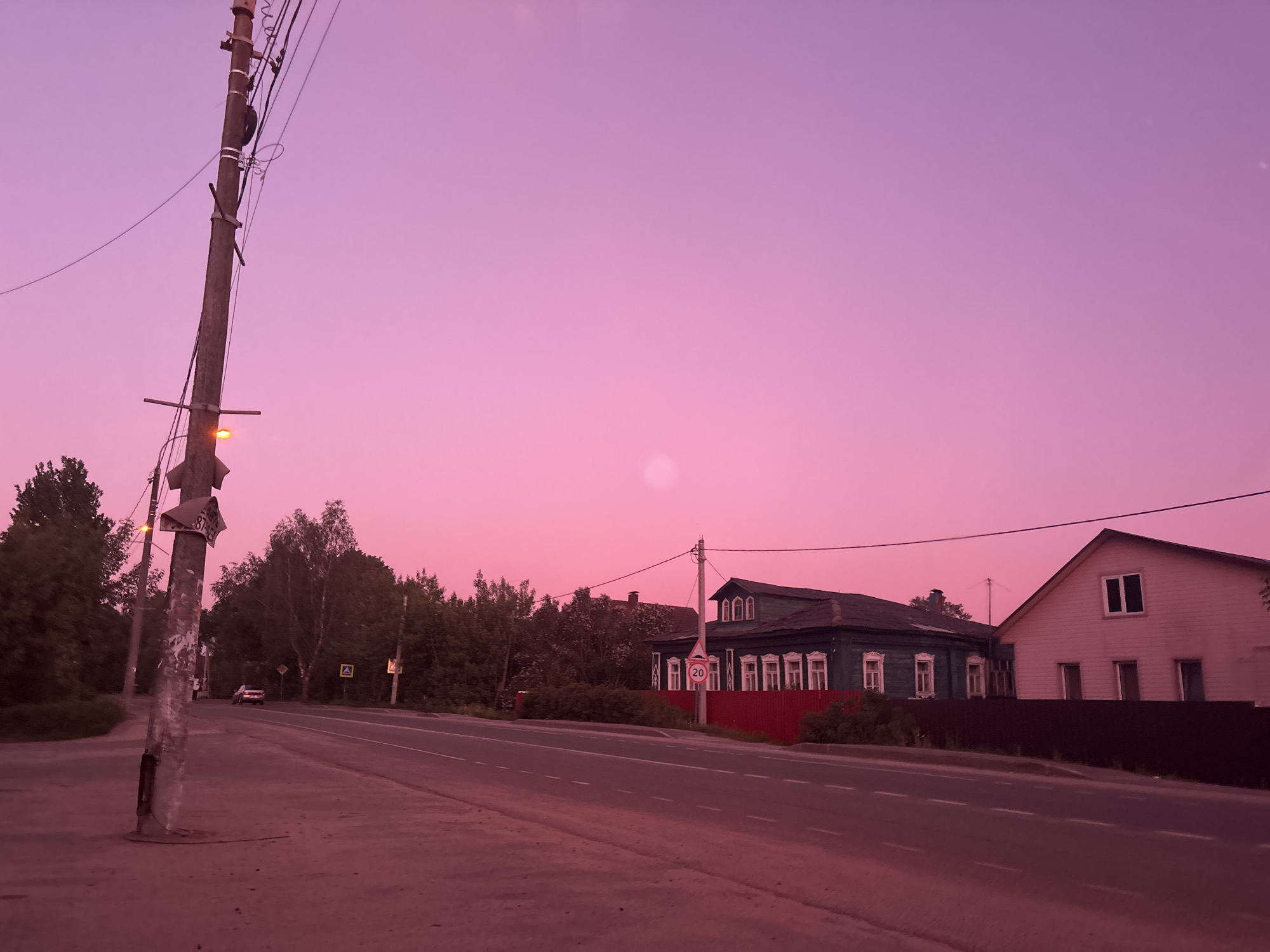 A street scene in Zaprudnya, Russia, at dusk or dawn, bathed in pink light. A two-lane road runs horizontally through the image, with sidewalks on either side. A utility pole with wires and a streetlight stands on the left sidewalk. Across the road, two houses are visible: a dark-green house with red fencing and a speed limit sign, and a light-colored house with dark fencing. Trees line the road behind the houses. The sky is uniformly pink with a bright spot. A car is traveling down the road in the distance.