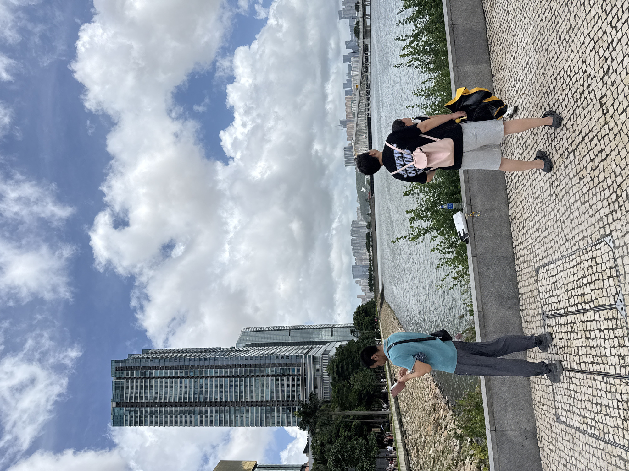 In Macau, China, on a cloudy day, two individuals stand on a paved walkway overlooking a body of water. The person on the left, facing away from the camera, is wearing a blue t-shirt and grey pants, holding a book or tablet. A small bag is slung across their shoulder. The person on the right, also facing away, is wearing a black t-shirt with a graphic print and grey shorts, carrying a pink backpack. They are holding a yellow and black object, possibly an umbrella or bag. In the background, several modern high-rise buildings dominate the skyline, with a bridge spanning the water. Trees and a rocky embankment line the water's edge. The sky is filled with dramatic, billowy clouds.