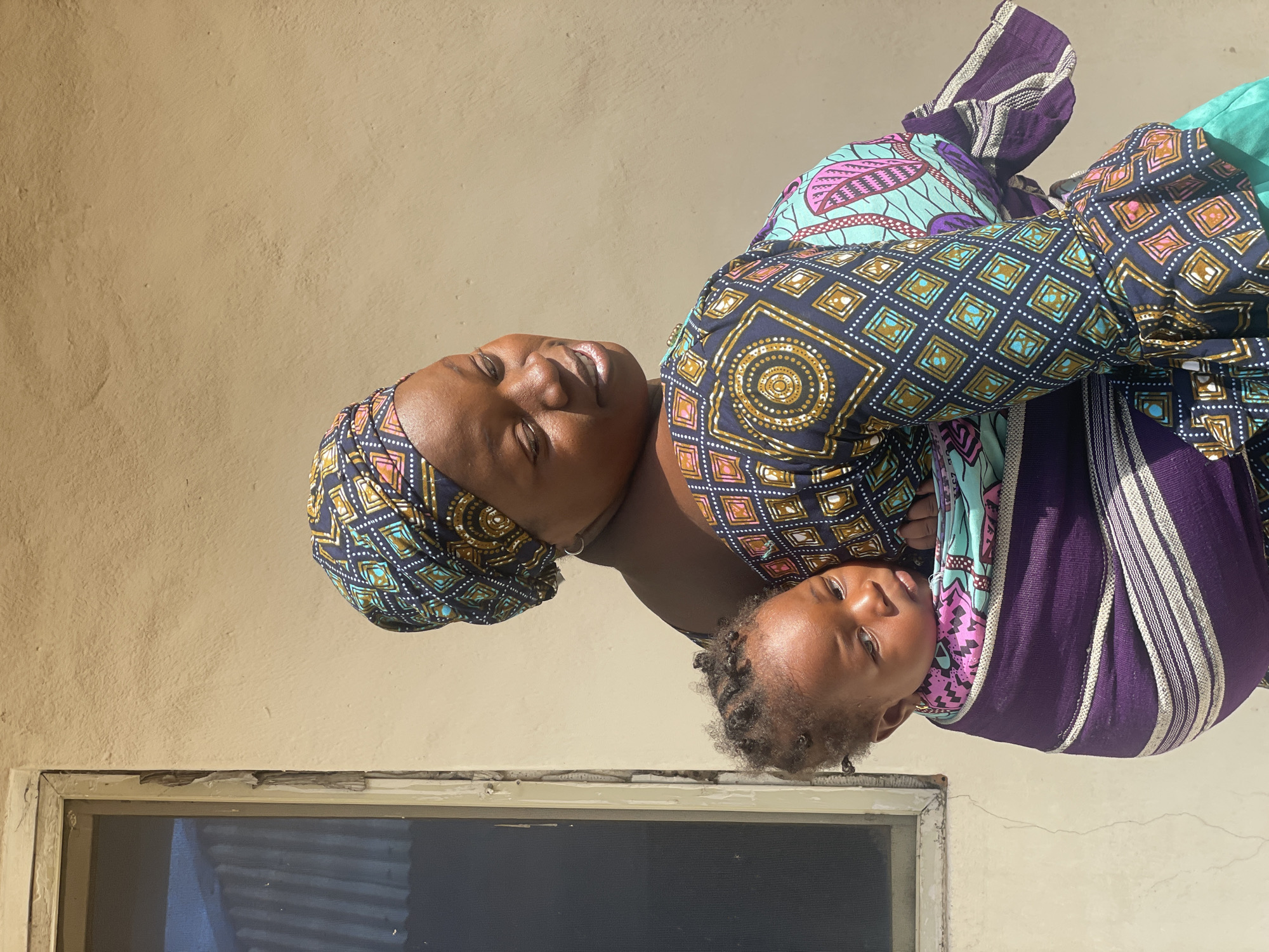 A woman holds a baby in a carrier in front of a light tan wall. The woman is wearing a colorful patterned head wrap and dress. The baby is secured in a purple and white striped carrier. The location is likely in Lokoja, Nigeria. There is a window in the bottom left corner of the frame.