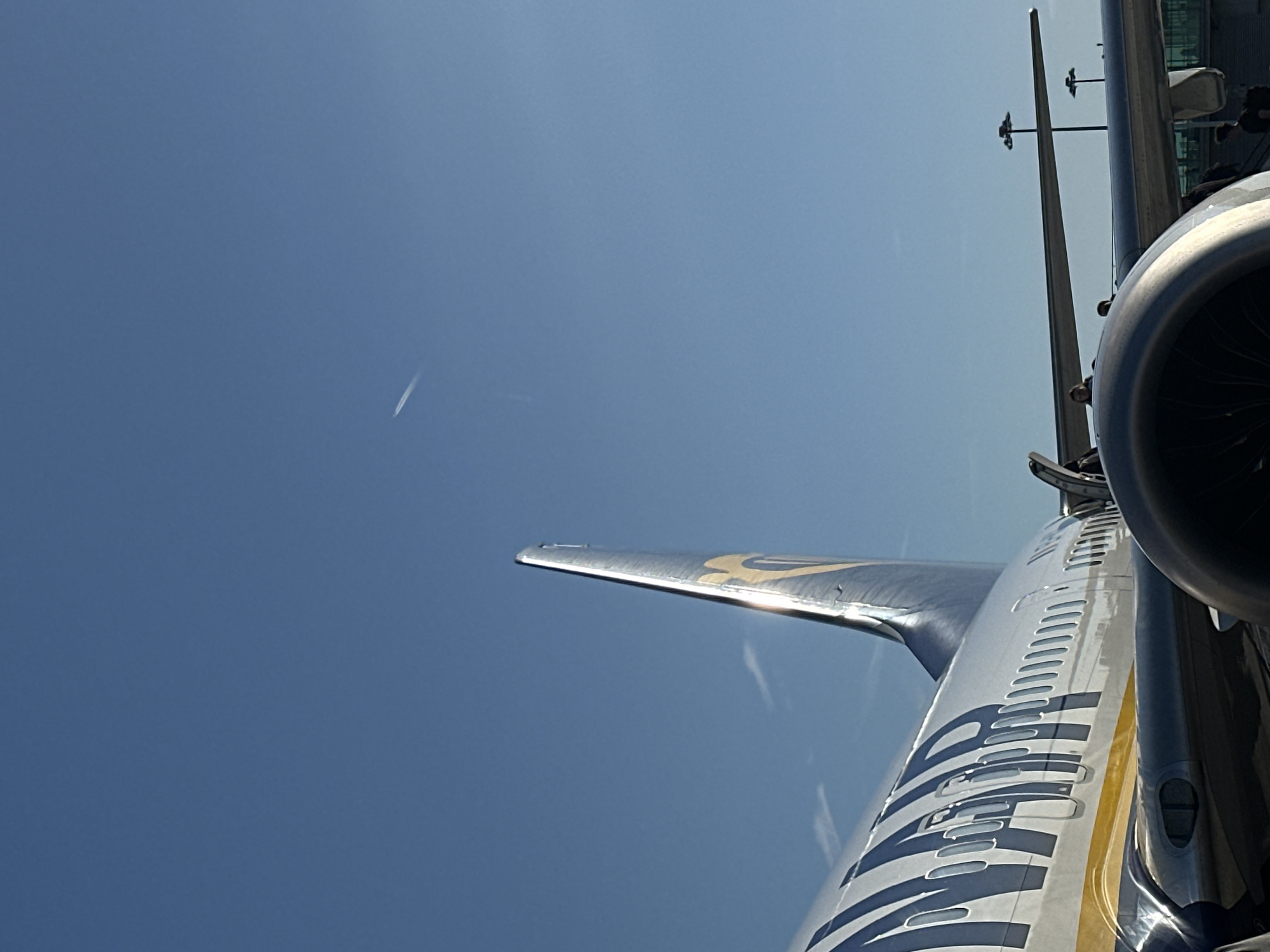 The image shows a partial view of a Ryanair airplane against a clear blue sky with a contrail visible in the upper left. The aircraft's wing with the Ryanair logo is prominent, as is part of an engine nacelle. In the background, a metal structure resembling an airport gate is visible. The location is likely an airport, and given Ryanair's extensive European network, this is possibly in Uttlesford, United Kingdom if the photo was taken there, or any airport within its network. The scene depicts a stationary airplane, possibly during boarding or disembarkation. People are visible in the background near the airport gate structure.