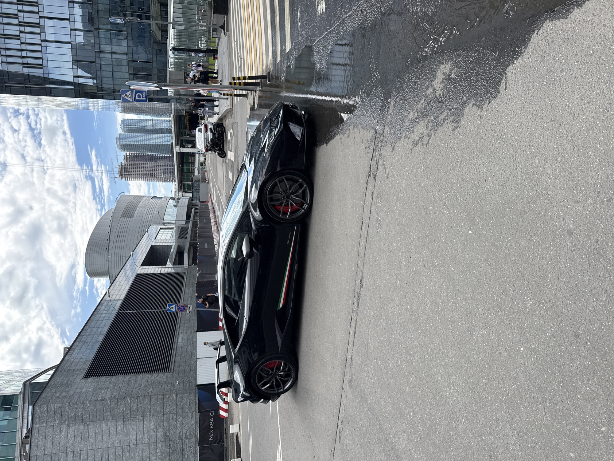 The image shows a black Lamborghini parked on a gray asphalt road with puddles of water in Khimki, Russia. In the background are modern high-rise buildings and a pedestrian crosswalk with people walking. A parked white motorcycle is also visible. The car features red brake calipers and an Italian flag stripe. Signs in Cyrillic are present.