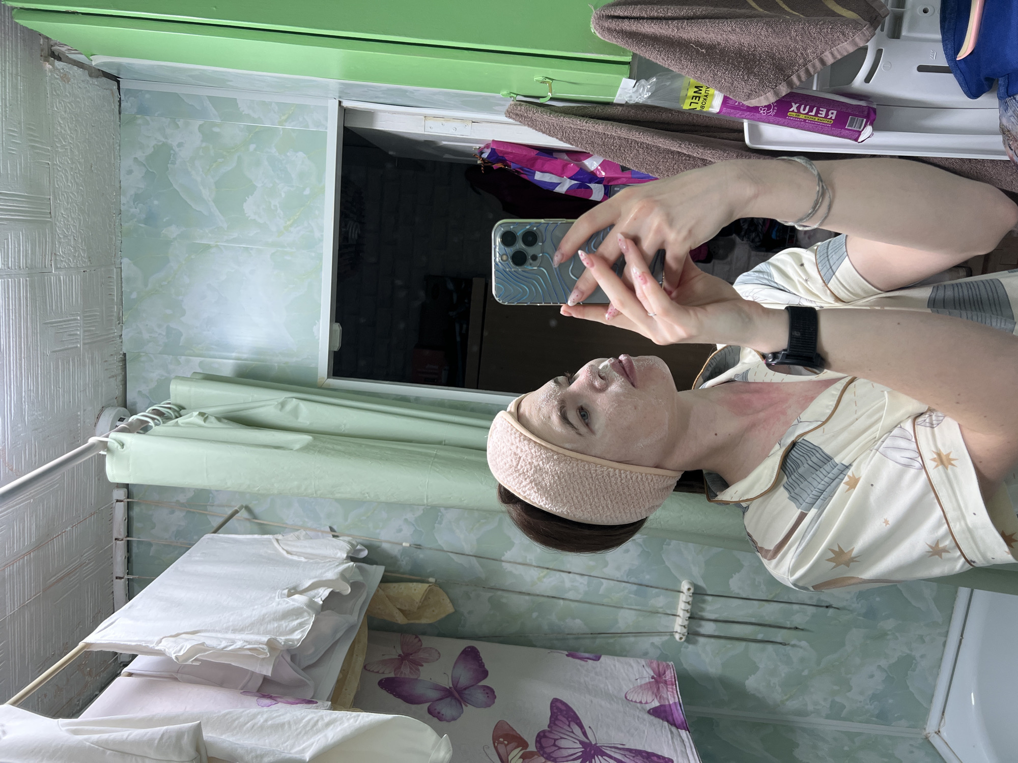A person takes a mirror selfie in what appears to be a functional room, possibly a bathroom or utility area, in Chaikovsky, Russia. The main subject is a woman with her hair pulled back by a light pink textured headband, and her face is covered in a white skincare product or mask. She is wearing light-colored pajamas adorned with star and abstract patterns. A black smartwatch is visible on her left wrist, and a silver bracelet is on her right arm. Her nails are manicured with a light polish. There is noticeable redness on her neck and upper chest.
She is holding a smartphone, an iPhone with a distinct patterned case (blue and silver wave-like design), to capture her reflection. The room's walls are covered in different styles: one section features light greenish-white panels with a marble-like pattern, while another has textured white tiles. A vibrant green frame or shelf is visible at the top.
In the background, a drying rack holds various items, including white t-shirts and a light green fabric. Below this, a fabric with a purple butterfly pattern is draped. To the right, brown towels hang from a hook, and a white tray holds a purple package with the word "RELUX" visible backward in the reflection. The overall lighting suggests an indoor setting, and the presence of the drying rack and skincare routine elements points to a practical, everyday environment.