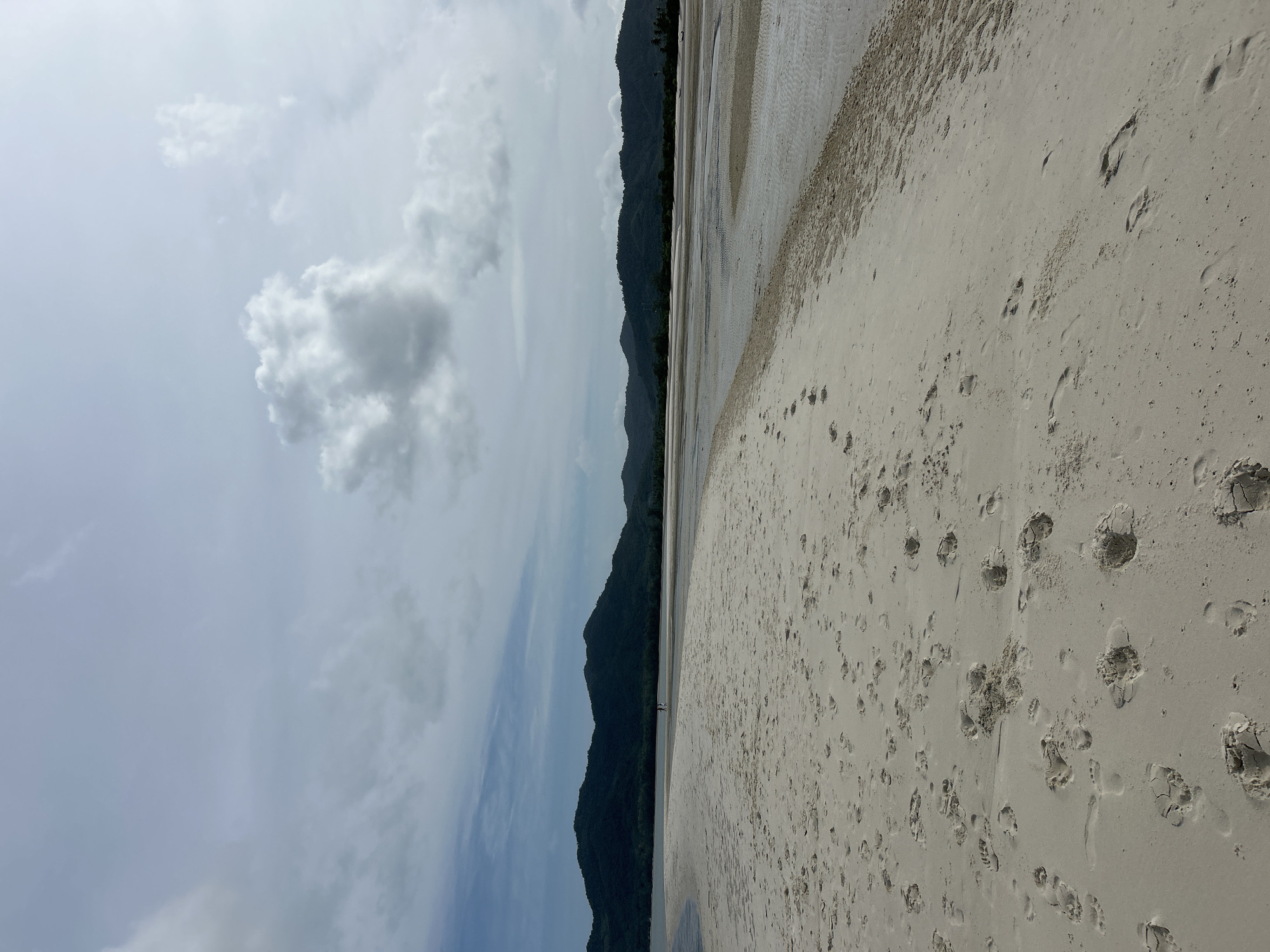 The image depicts a wide, sandy beach with numerous footprints visible in the foreground. In the mid-ground, two figures are walking away from the camera on the sand, which is adjacent to a body of shallow water. The background features a line of dark green, hilly terrain, likely mountains, under a cloudy sky. The scene appears to be a natural landscape, possibly a coastal area in Ko Yao, Thailand. The overall impression is one of a serene, natural environment with signs of human presence.