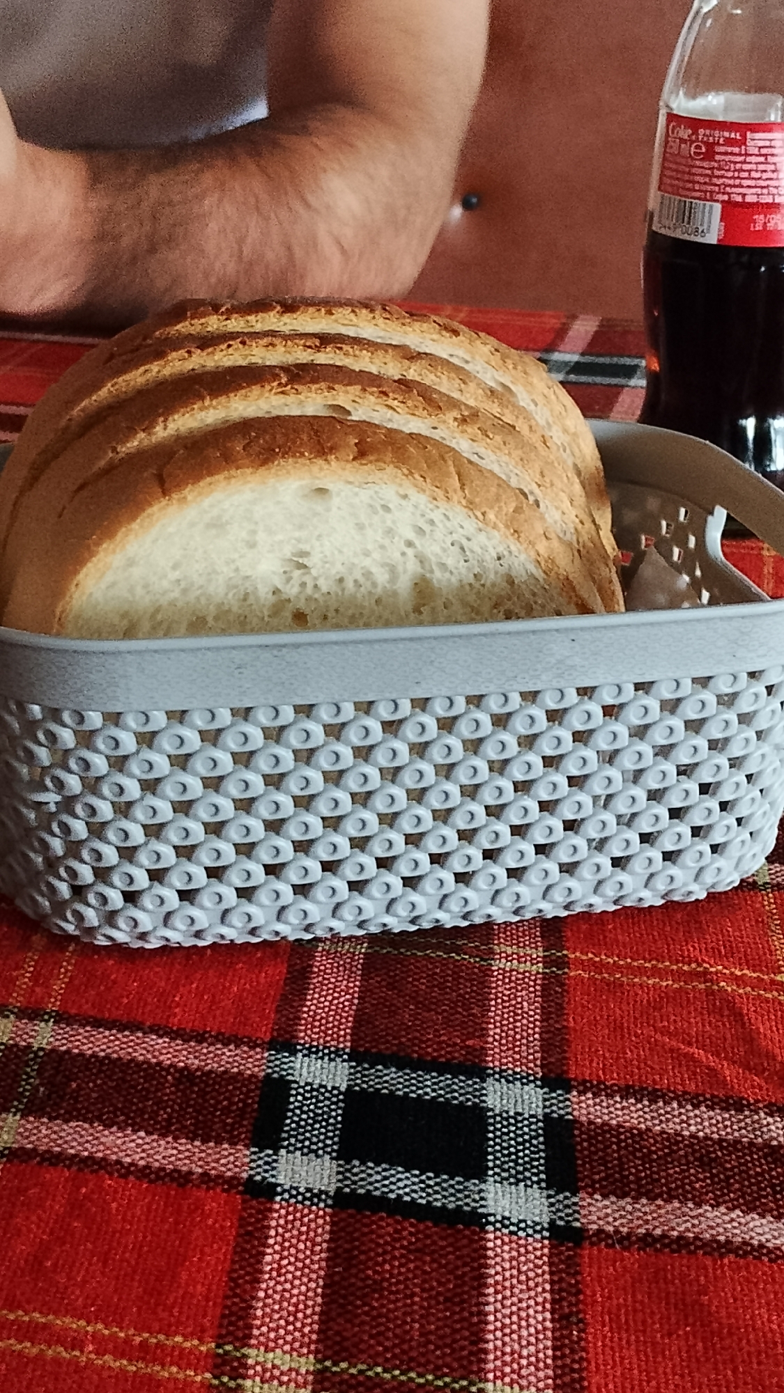 A close-up shot shows a basket filled with sliced white bread. The basket is made of light gray plastic with a woven pattern of circles. The bread slices are stacked, with their golden-brown crusts visible on top. In the background, partially visible, is a person's arm and shoulder, with dark hair and a grey t-shirt. To the right of the bread, a dark brown glass bottle with a red and white label, appearing to be a Coca-Cola bottle, is visible. The scene is set on a red and black plaid tablecloth. The image does not provide any information about the city of Pevtsite, Bulgaria.