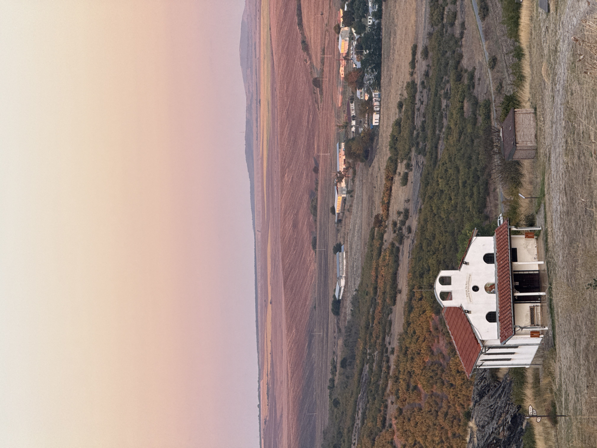 The image depicts an outdoor scene in the city of Rusokastro, Bulgaria, at dusk. In the foreground, a small white church with a red tile roof stands prominently on a grassy knoll. The church has arched windows and a covered entrance with a bench. To the right of the church, a small, wooden shed-like structure is visible.
The middle ground features a landscape of rolling hills covered in dry grass and low-lying vegetation, with scattered trees. Further in the distance, a cluster of buildings, likely part of a village, can be seen. These buildings are varied in size and architectural style, with some having red tile roofs.
The background is dominated by vast agricultural fields, appearing to be recently harvested or plowed, with distinct patterns of tilled soil in shades of reddish-brown and muted yellow. A gentle slope of hills forms the horizon, under a sky displaying a gradient of soft pastel colors, transitioning from pale yellow and peach at the top to a light pink and purple towards the horizon, indicating the setting sun. The overall atmosphere is serene and rural.