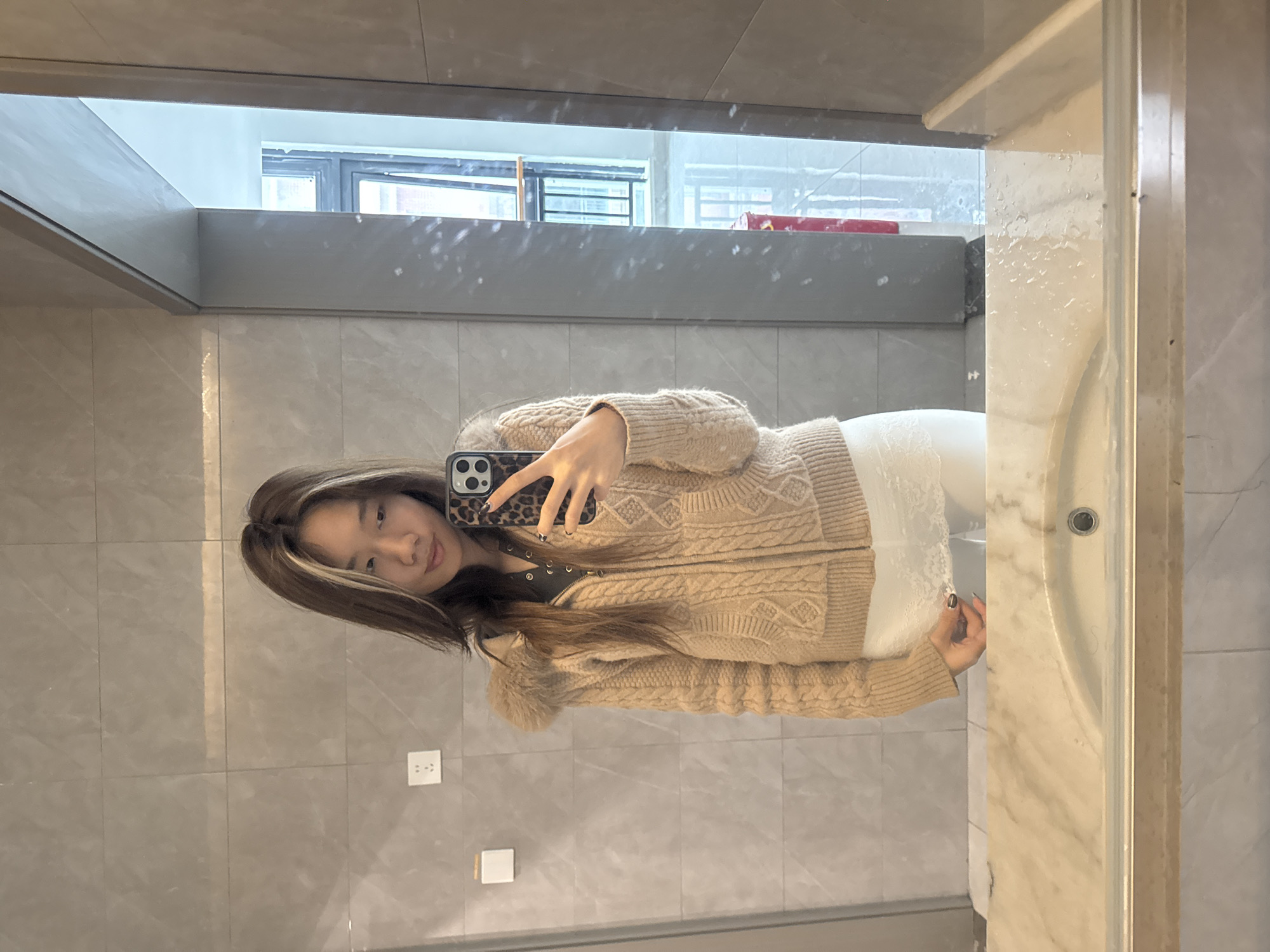 A woman is taking a selfie in a bathroom in Nan'an District, China. The bathroom has grey tiled walls and a marble-like countertop with a sink on the right. The woman is wearing a beige cardigan and white pants. A window can be seen in the background. She is holding a phone with a leopard-print case.