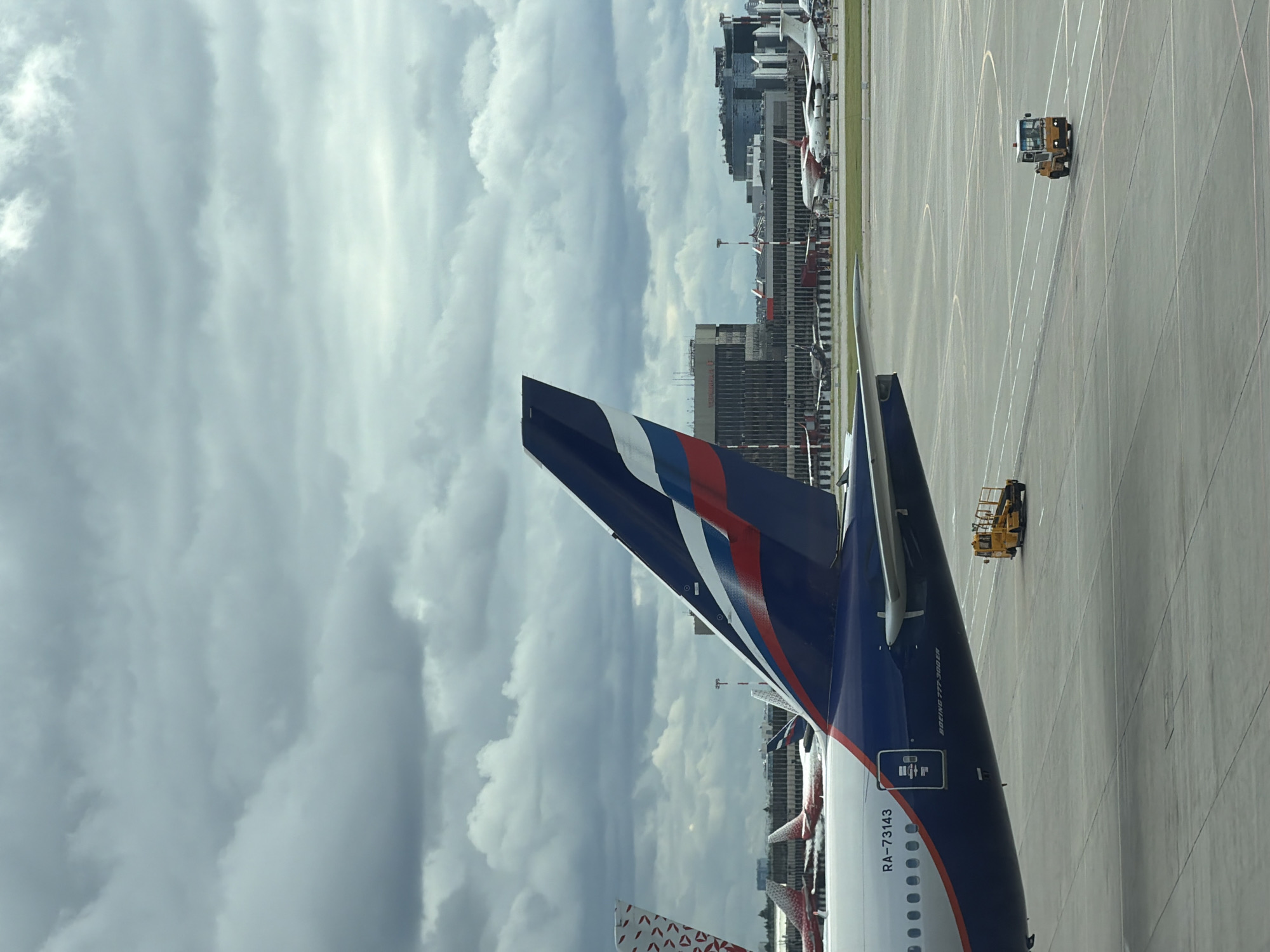 An eye-level, medium shot captures an airport tarmac under a cloudy sky. In the foreground, the tail section of a large airplane, painted in dark blue with white and red stripes, dominates the left side of the frame. Visible text on the fuselage reads "RA-73143" and "BOEING 777-300ER". To the right, a yellow airport vehicle, resembling a tow tractor, is parked on the tarmac. In the midground, other airplanes are visible, as are various airport buildings with glass facades. Another yellow airport vehicle is moving on the tarmac in the distance, on the right side of the frame. The scene is a wide, open airport environment, suggesting operations under potentially inclement weather. The image was taken at Sheremetyevo International Airport in Khimki, Russia.