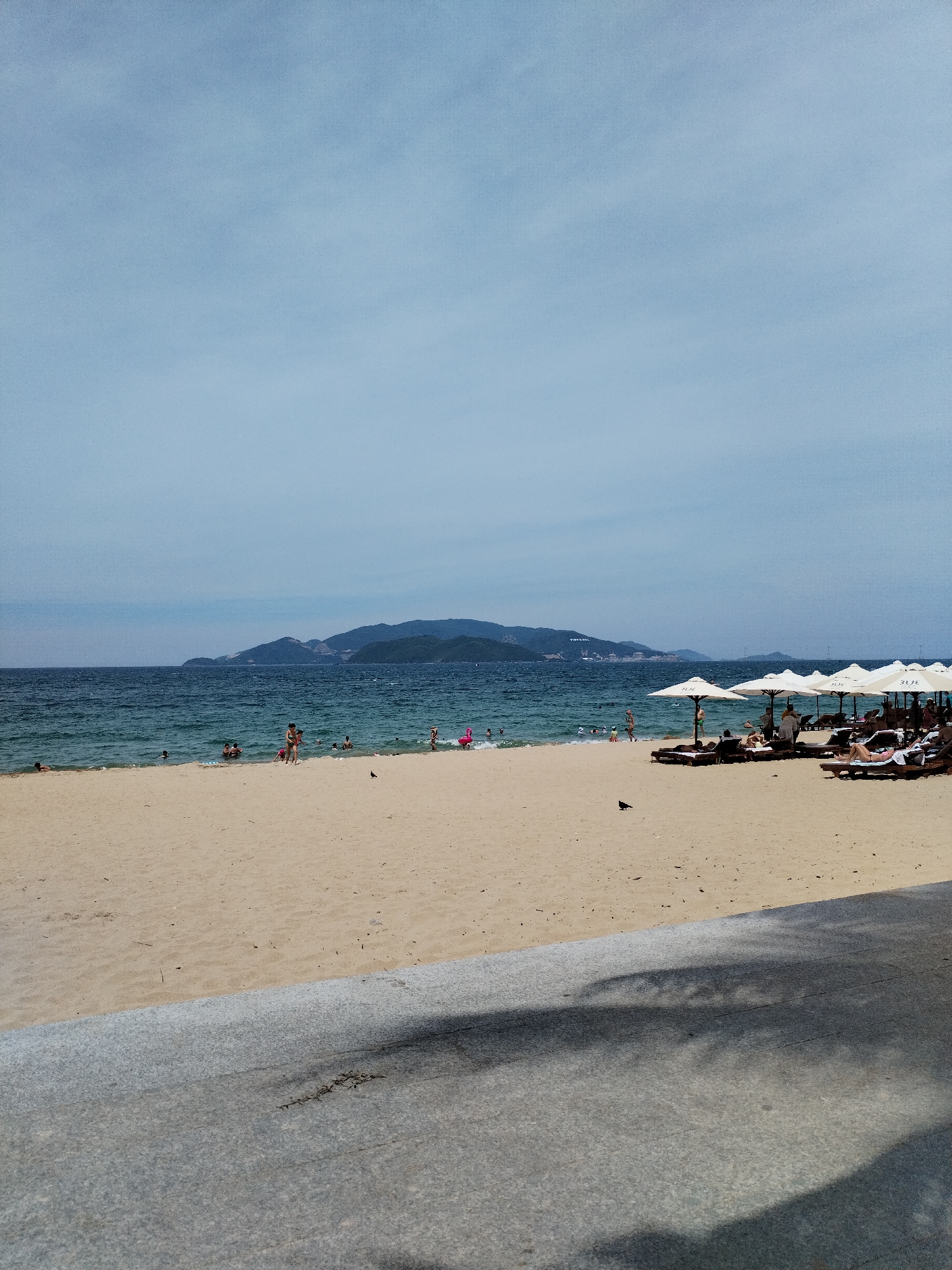 This is a wide shot of a beach in Nha Trang, Vietnam, on a clear day. In the foreground, a concrete walkway casts shadows of palm trees onto its surface. Beyond the walkway is a wide expanse of light-brown sand. The ocean, a deep blue, stretches to the horizon, where a hazy, mountainous coastline is visible.
Scattered across the sand, closer to the water, are several beach umbrellas providing shade for lounge chairs. Some chairs are occupied by people relaxing. A small number of individuals are visible in the shallow water, either swimming or wading. A few people are also walking along the shoreline.
The sky above is a pale blue with some wispy clouds. A lone dark bird is perched on the sand in the middle ground. A pink inflatable flamingo can be seen near the water's edge on the left side of the image. The scene depicts a typical day at a popular beach resort.