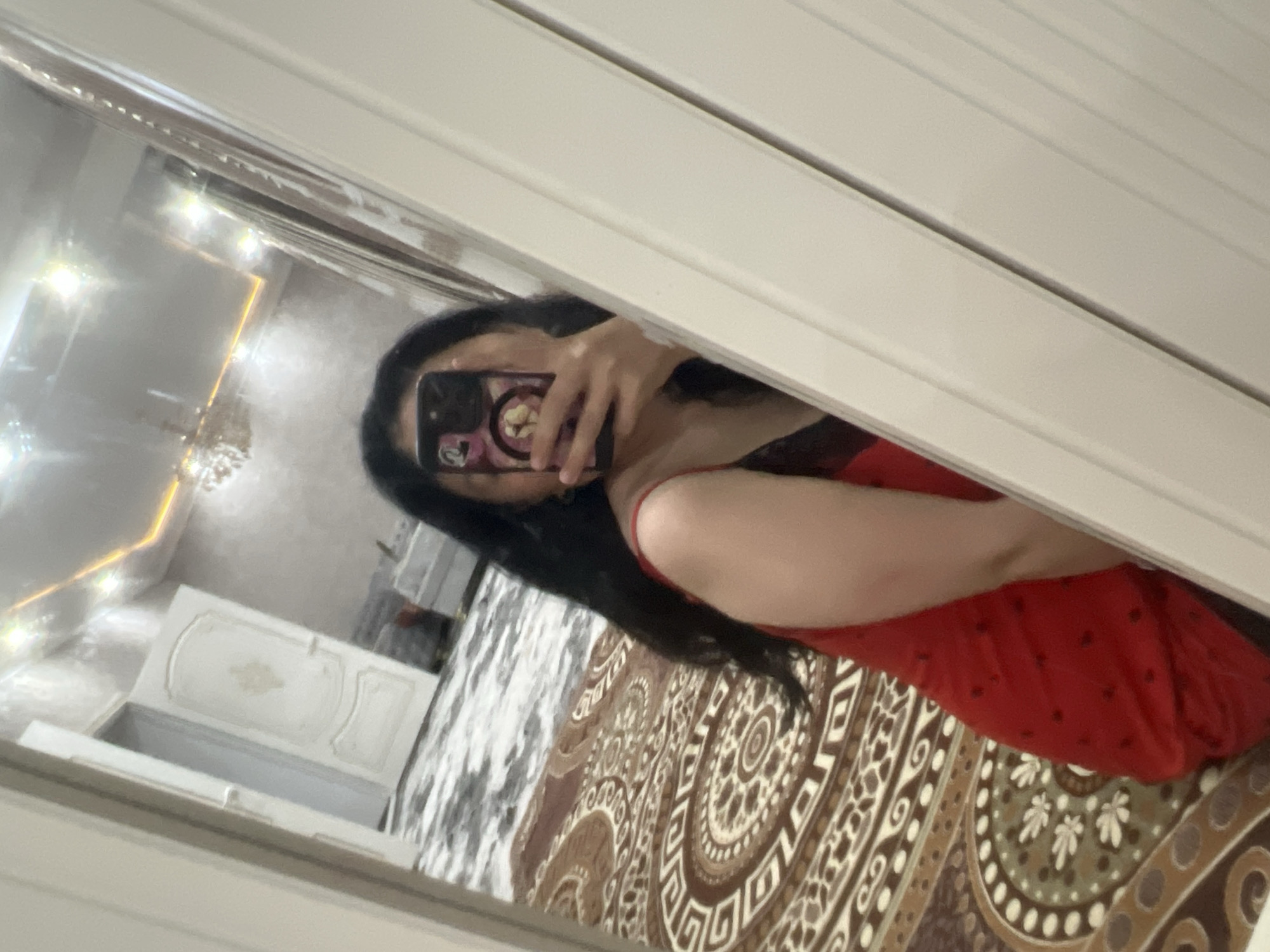 The image is a vertical, slightly blurred selfie taken in a room. A person with long, dark hair is visible, holding a phone up to take the photo. They are wearing a red garment with thin straps. The background, reflected in a mirror, shows a room with a patterned carpet, a white door, and a ceiling with ornate lighting. The location is not specified in the image itself, so it cannot be determined to be in Kara-Dıykan, Kyrgyzstan.