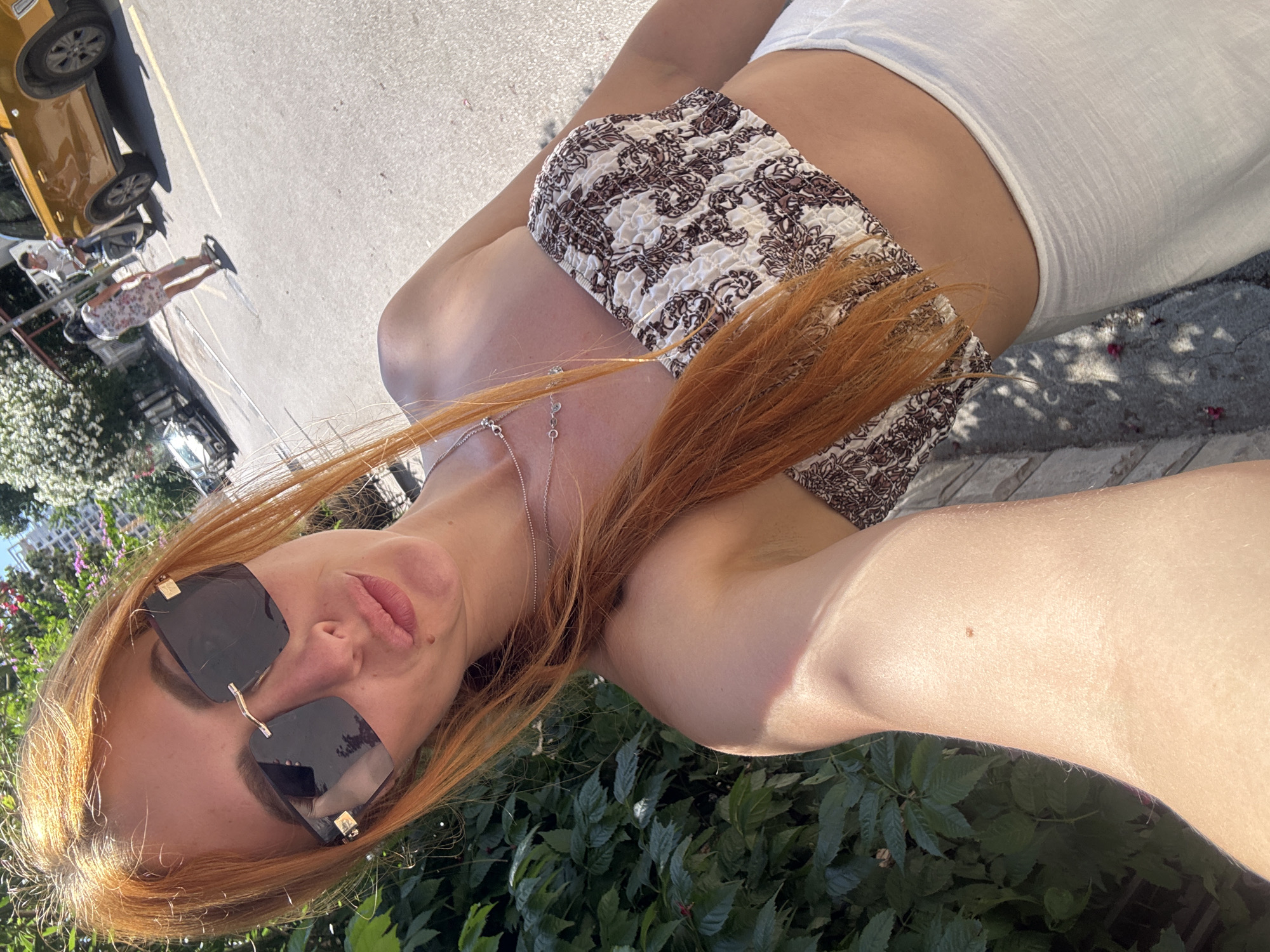 A woman with long, reddish-orange hair and dark, rimless sunglasses with gold accents poses for a selfie on a sunny day in Budva, Montenegro. She is making a slight pouting expression, with a small brown mole visible below her lower lip on the right side. Her hair is draped over her left shoulder, and she wears multiple delicate silver chain necklaces. Her attire consists of a strapless, ruched crop top featuring a white background with brown and black paisley-like patterns, paired with loose-fitting white bottoms. Her right arm, visible in the foreground, exhibits sun-kissed skin with light arm hair and a small brown freckle or mole on the upper arm.
The background shows a paved street or road under bright daylight, with visible yellow lane markings. To the woman's immediate left, dense green foliage with scattered small red flowers is present. On the street, a woman in a light-colored floral dress walks away from the camera, carrying a light bag. Further down the street, a man is partially visible, positioned near a distinctive mustard-yellow SUV parked on the right side of the road, facing away. Another light-colored car is parked further down on the left side. In the distance, white buildings and green trees are visible under a clear blue sky. Direct sunlight casts noticeable shadows across the scene.