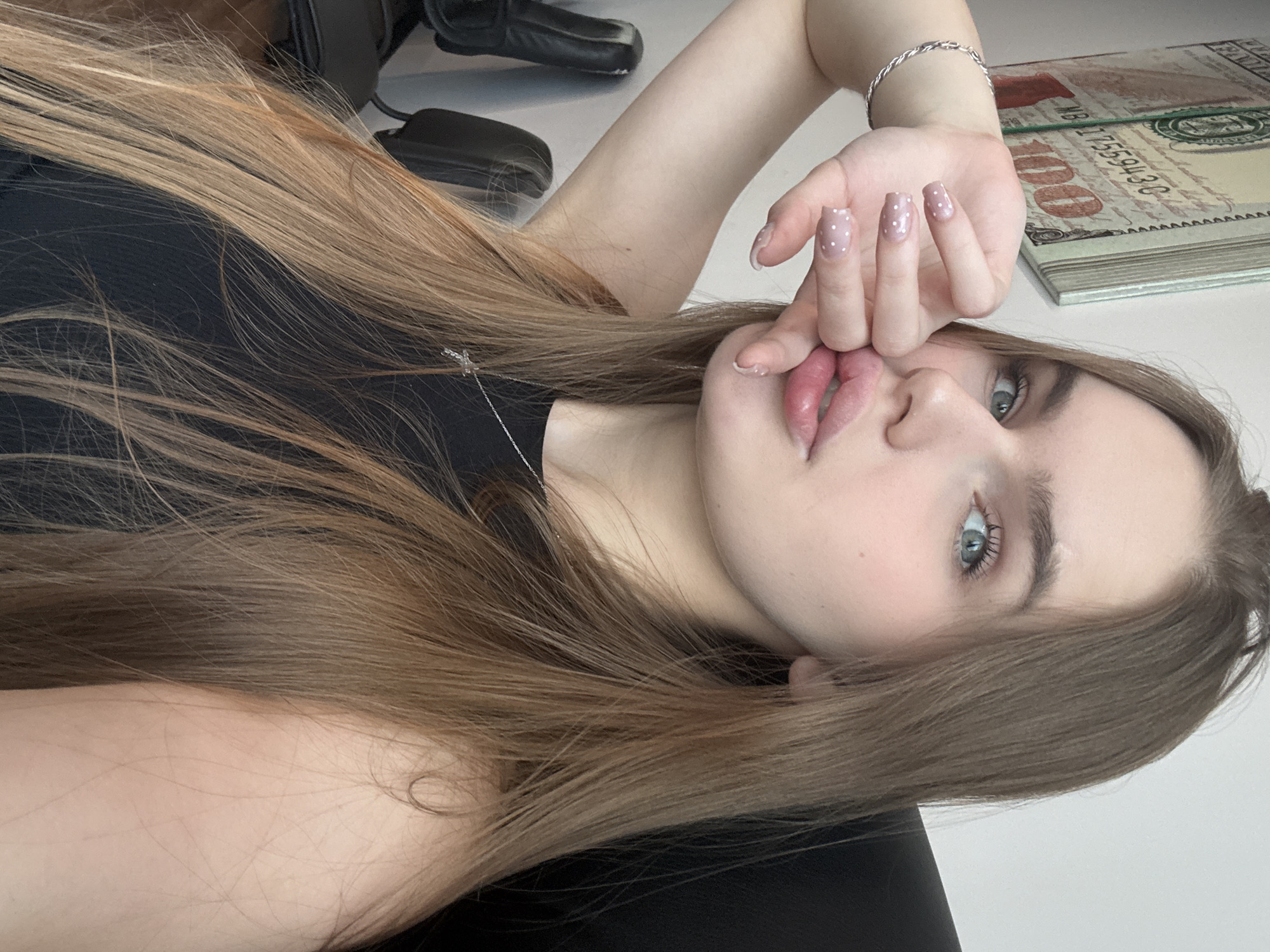 A young woman with long, light brown hair and blue eyes is pictured in a close-up, tilted shot. She is wearing a black top and has her right hand near her face, with her fingers lightly touching her lips. Her nails are painted a pale pink with white polka dots. A silver bracelet is visible on her wrist.
In the background, a stack of what appears to be prop money, resembling hundred-dollar bills, is resting on a white surface. A portion of a black office chair is also visible. The lighting suggests it is daytime, likely indoors. The overall mood is casual and relaxed.
No specific event or activity is clearly depicted beyond the subject posing for the photo. The location context of Dnipro, Ukraine, is provided but not evident from the visual cues within the image itself. There is visible text on the prop money, including "100" and what appear to be serial numbers and other imprints common on currency.